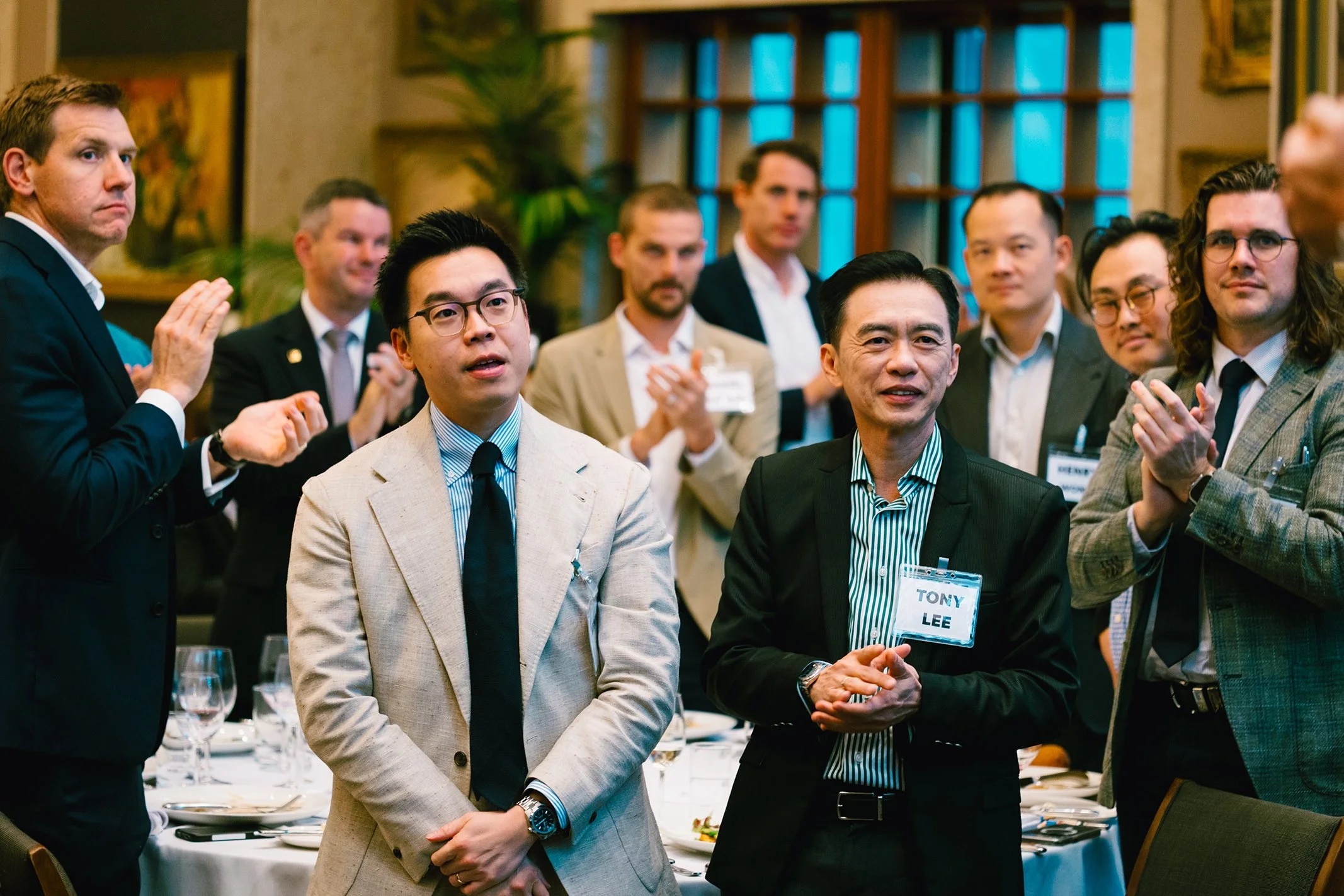 Group of men in formal attire at a professional event, some clapping, in a decorated room with table settings and large windows.