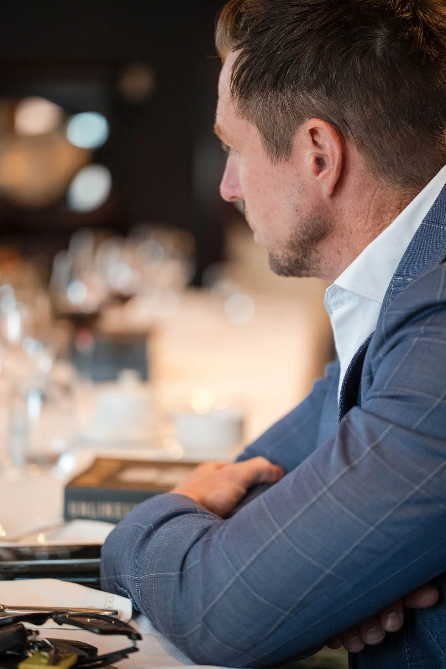 A man in a blue suit with glasses on the table in front of him, sitting with arms crossed, looking to the right in a restaurant or cafe.