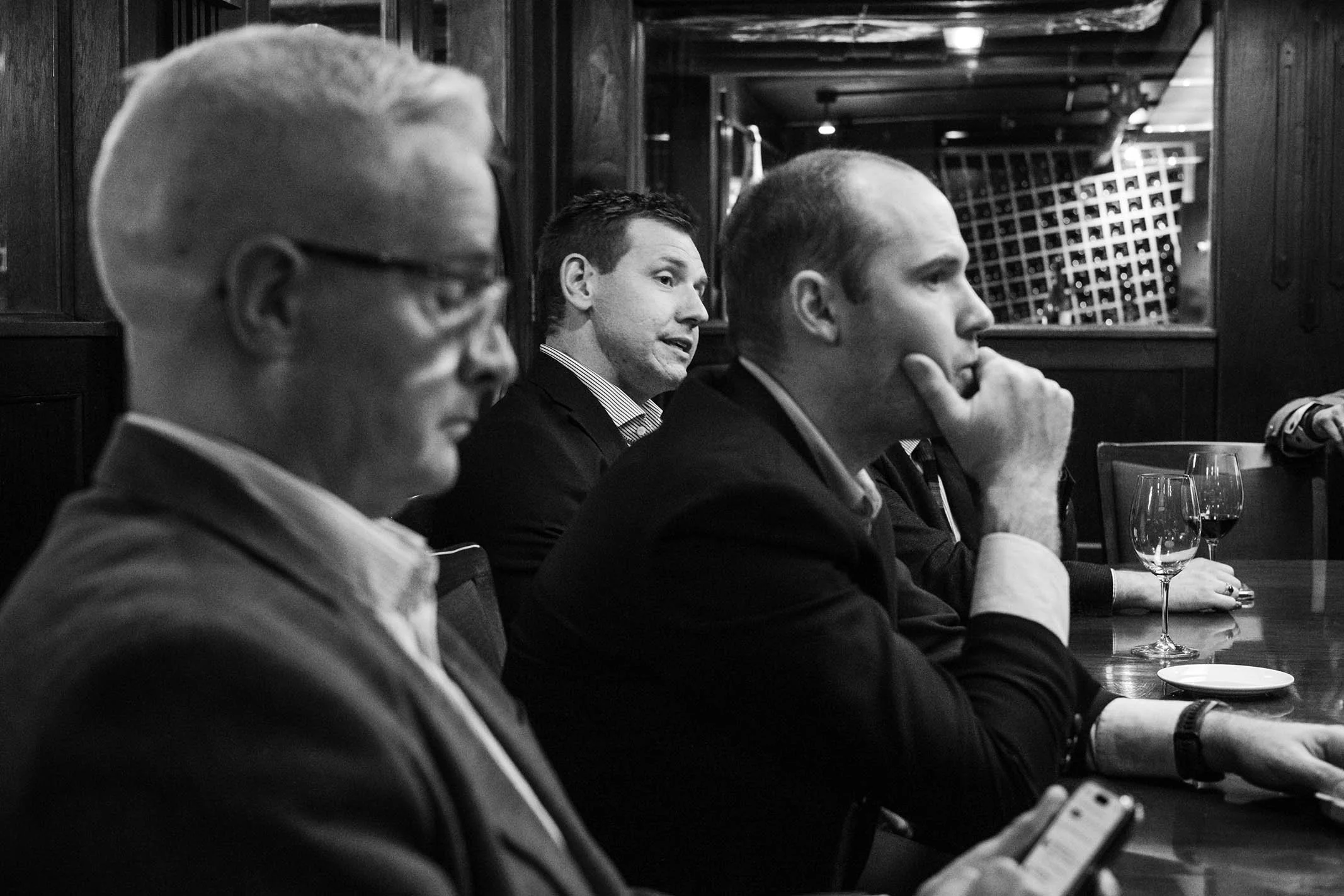 A black-and-white photo of four men sitting at a restaurant or bar, dressed in business attire, with glasses of wine on the table.