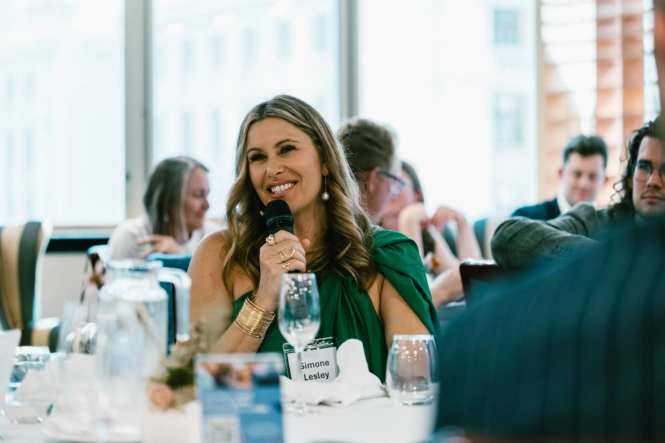 A woman in a green dress with long wavy hair, wearing pearl earrings and a gold bracelet, smiles and holds a microphone at a conference or event. She has a name tag that reads 'Simone Lesley' and is seated at a table with glasses of water and a flora