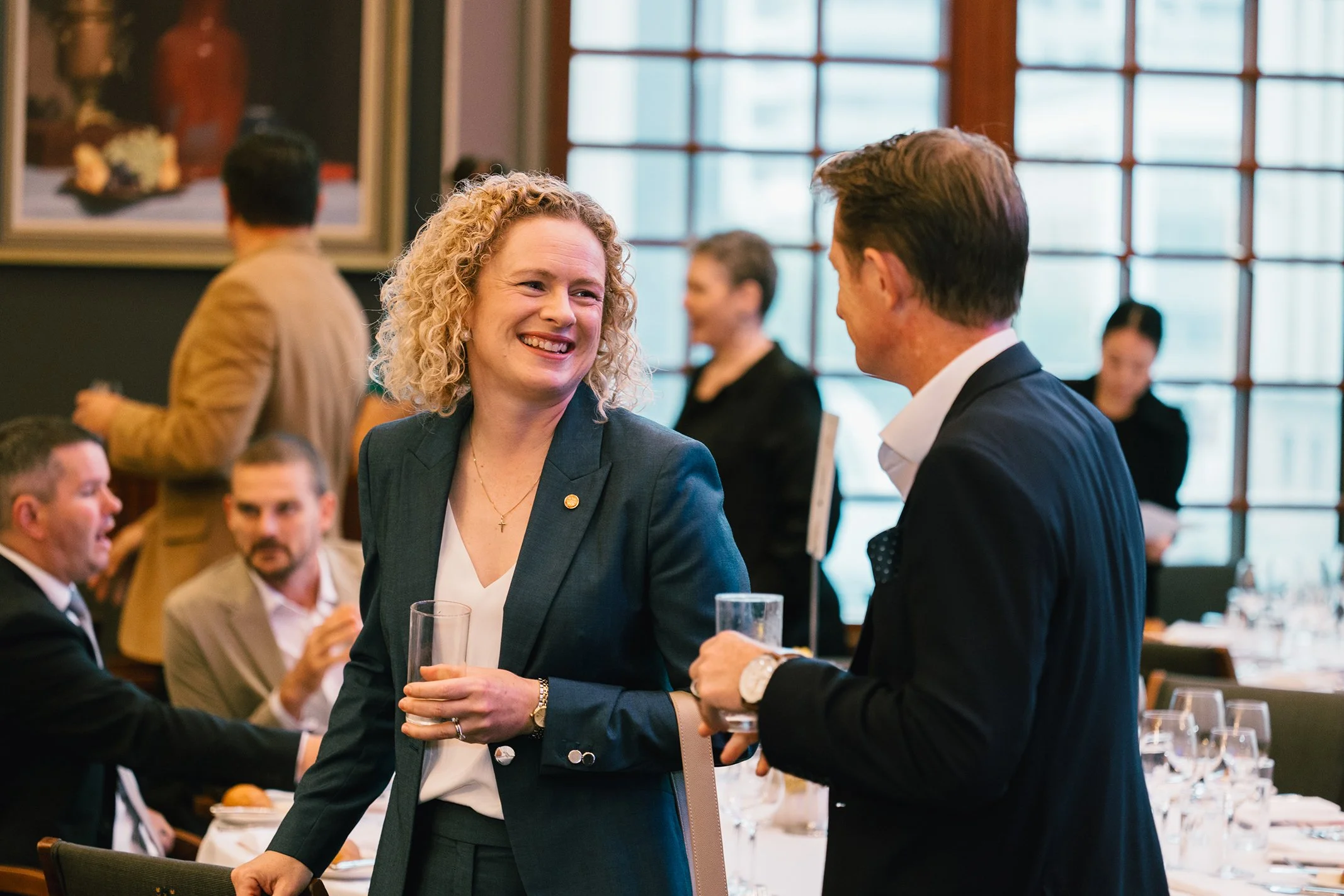 Two people in formal attire smiling and talking at a social event, with other guests in the background, in a well-lit room with large windows and a table set for dining.