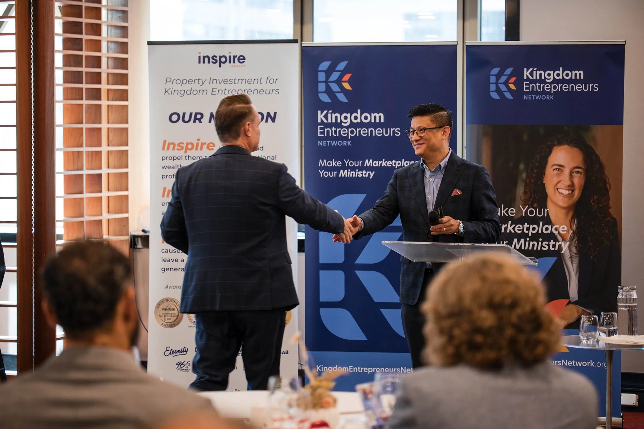 Two men shaking hands at a professional event, standing in front of banners that read 'Kingdom Entrepreneurs' and 'Inspire Property Investment for Kingdom Entrepreneurs' with an audience seated in front.