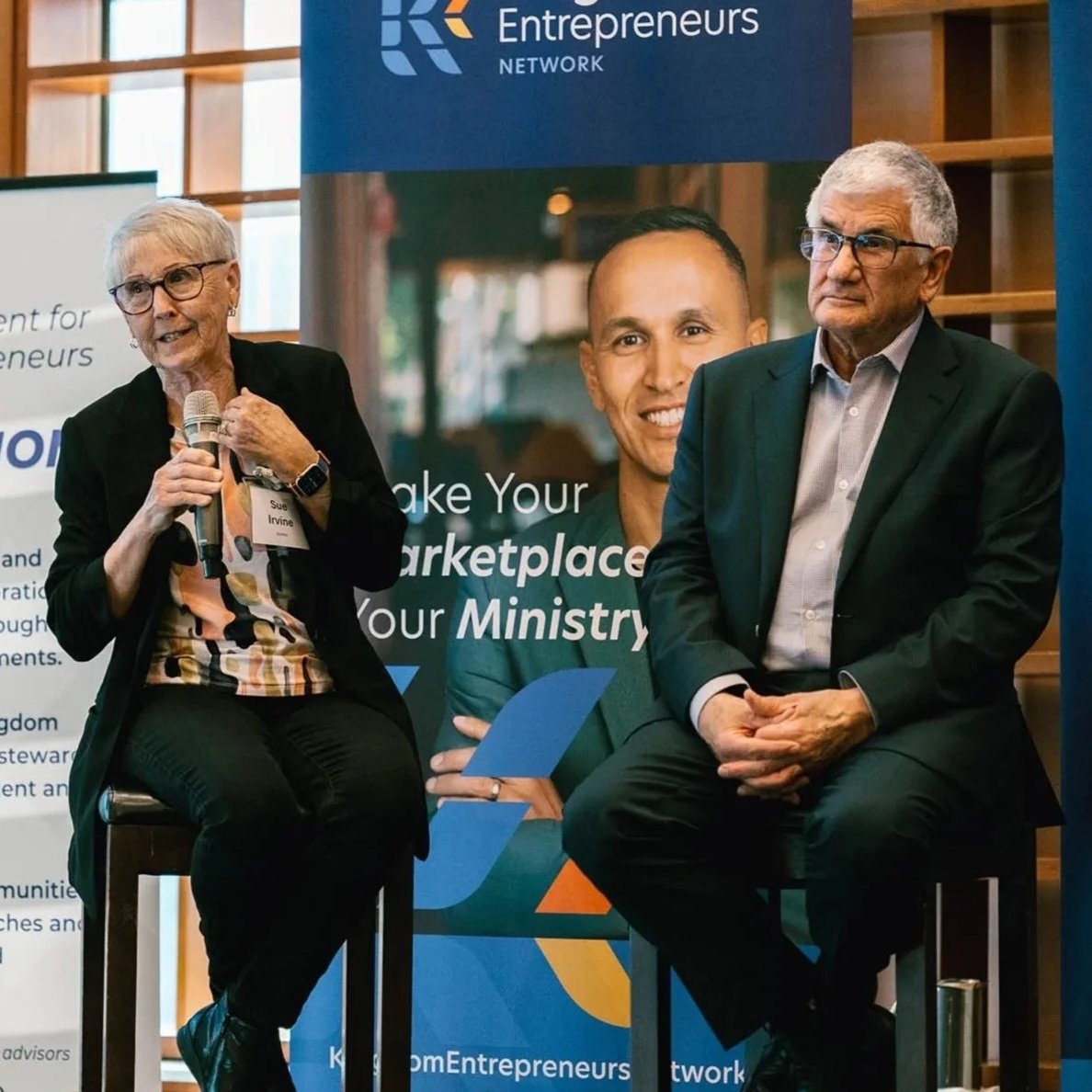 An older woman and an older man are seated on chairs at a panel discussion or event. The woman is holding a microphone and appears to be speaking, while the man sits with his hands clasped in his lap. They are in front of a large banner with the logos and branding of an entrepreneurship network, with text encouraging making a marketplace for a ministry. The woman has short gray hair, glasses, and is wearing a black jacket and a colorful shirt, while the man has gray hair, glasses, and is dressed in a dark suit with a light shirt.