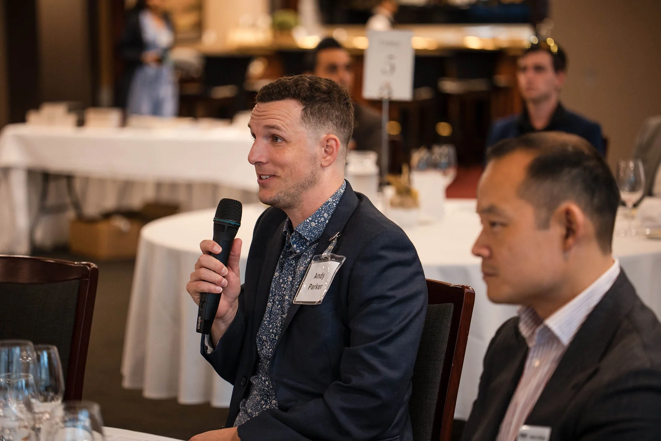 Man in dark suit holding a microphone and speaking at a round table in a formal event or conference setting, with other attendees seated nearby.