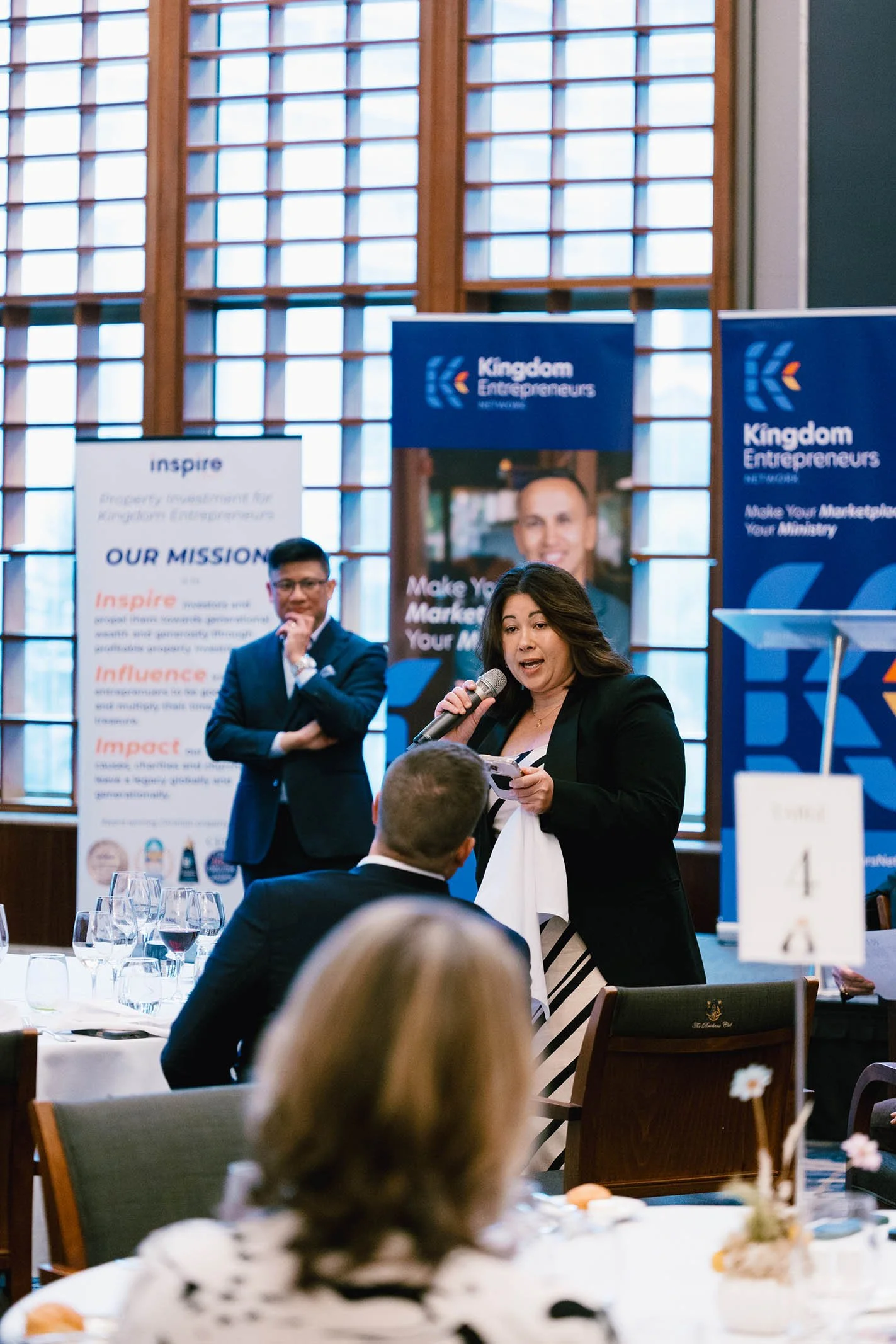 Woman speaking into microphone at a banquet event with attendees seated at tables, promotional banners in the background reading 'Kingdom Entrepreneurs' and 'Make Your Marketing Your Ministry,' and large windows in the venue.