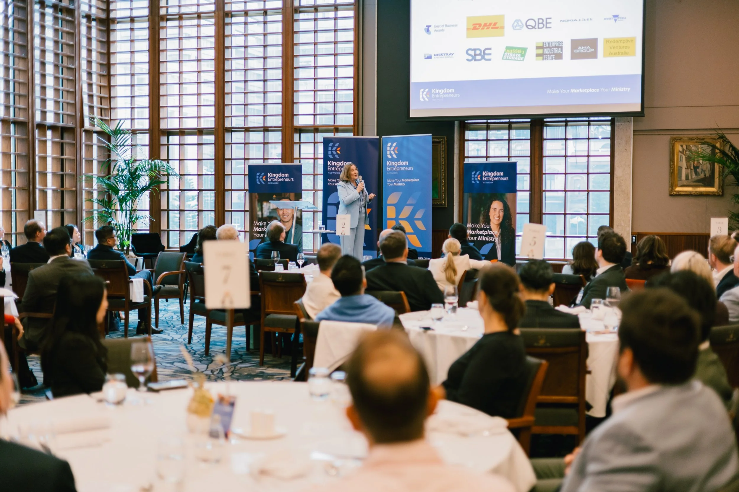 A woman presenter speaking at a business conference with an audience seated around round tables, large windows and banners behind her displaying the 'Kingdom Entrepreneurs' logo, and a screen showing sponsors' logos.