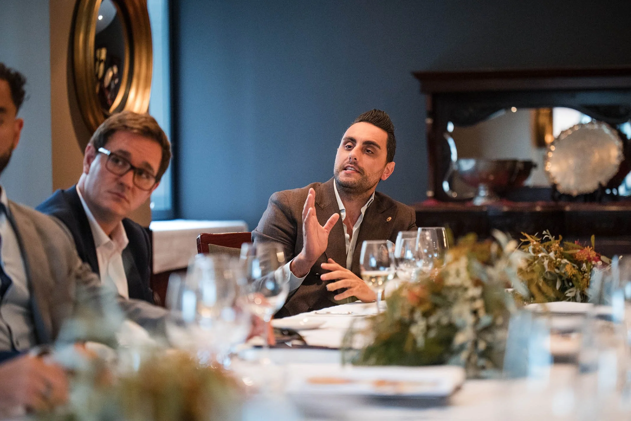 Man in brown blazer gesturing while speaking at a formal dinner event with other people seated around the table, decorated with flowers and glassware.