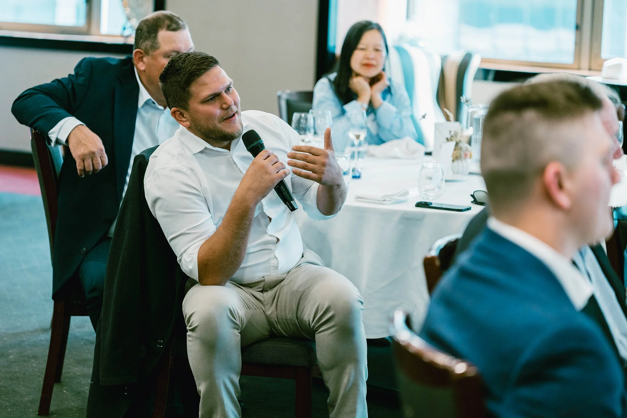 Man in white shirt speaking into a microphone during a formal event, with seated attendees and a woman in the background.