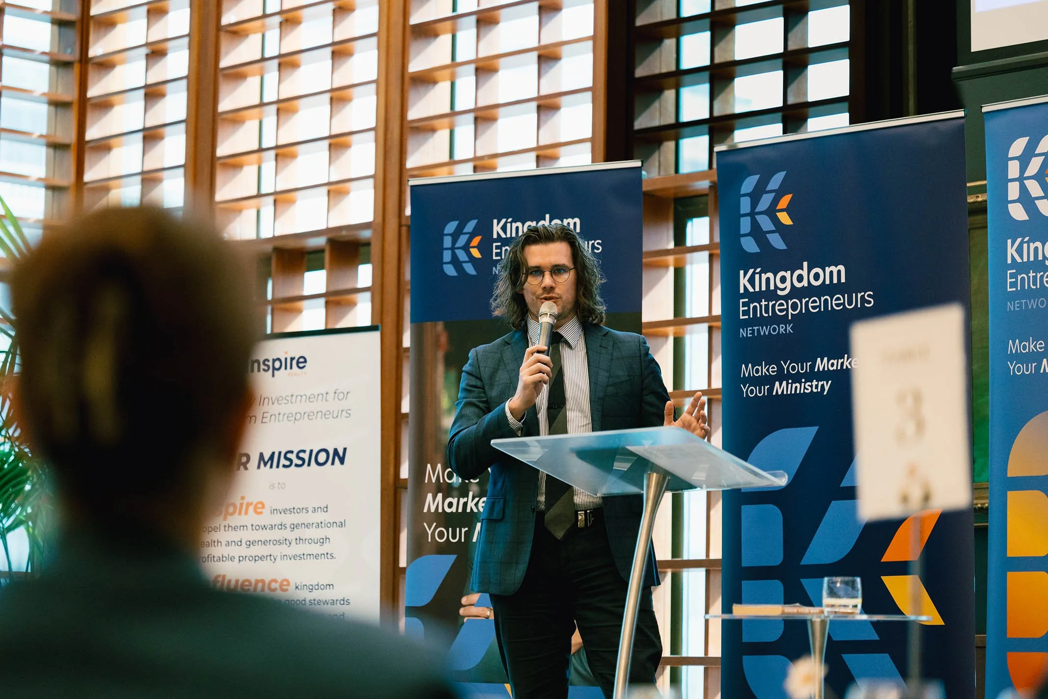 A man with long dark hair, wearing glasses and a suit, is speaking into a microphone at a presentation event for Kingdom Entrepreneurs Network. Behind him are blue banners with the network's logo and slogans.