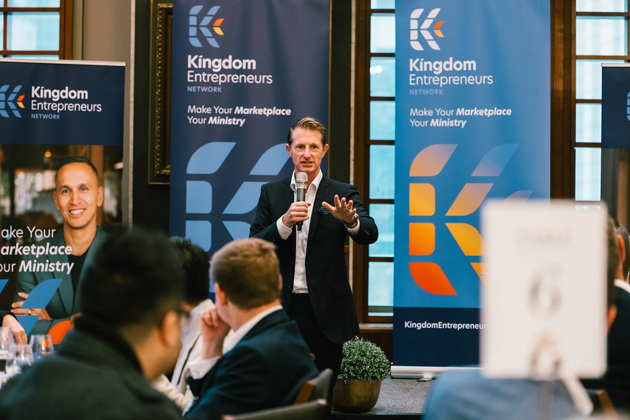 A man in a black suit and white shirt is holding a microphone and speaking to an audience at a conference. Behind him are two large banners with the logo and name "Kingdom Entrepreneurs Network" and the slogan "Make Your Marketplace Your Ministry." T