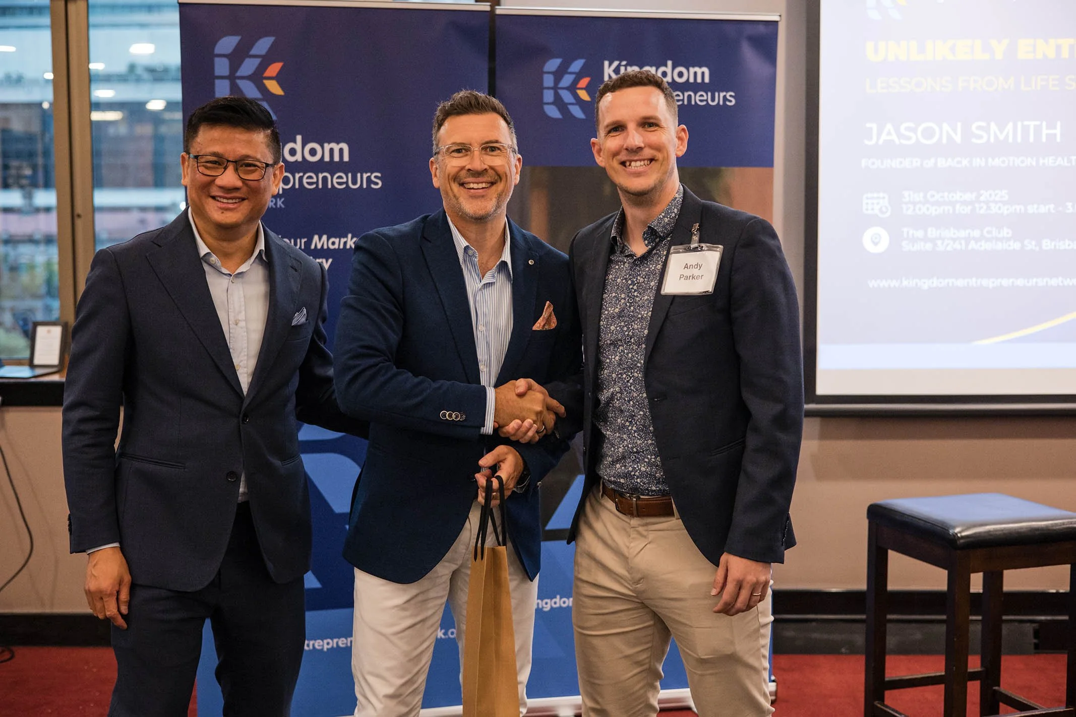 Three men in formal attire standing and smiling at an event, two shaking hands in front of a backdrop with the words "Kingdom Entrepreneurs Network" and a presentation screen on the right displaying event details and a person's name, Andy Parker.