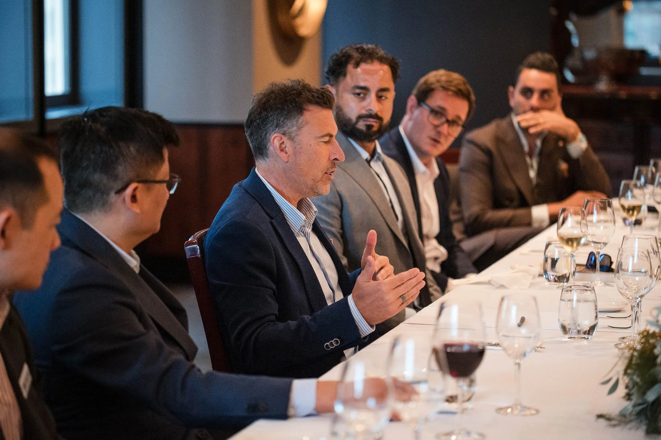 A group of men in formal business attire sitting around a conference table with wine glasses, engaging in discussion or listening attentively.