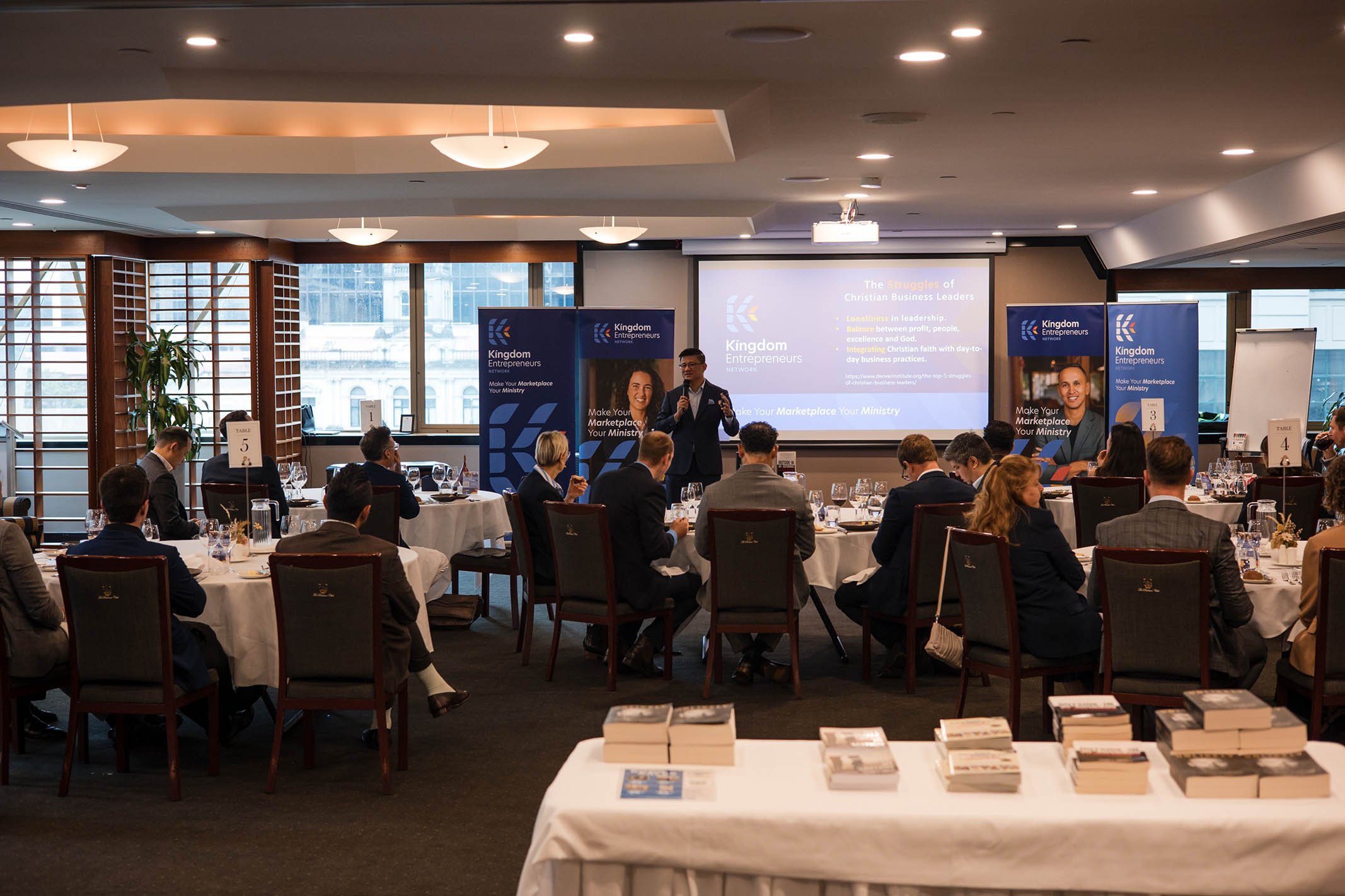 A business conference with a speaker presenting on stage at a round table event in a high-rise conference room. The room has large windows, white tablecloth-covered tables, and attendees in formal business attire listening to the presentation.