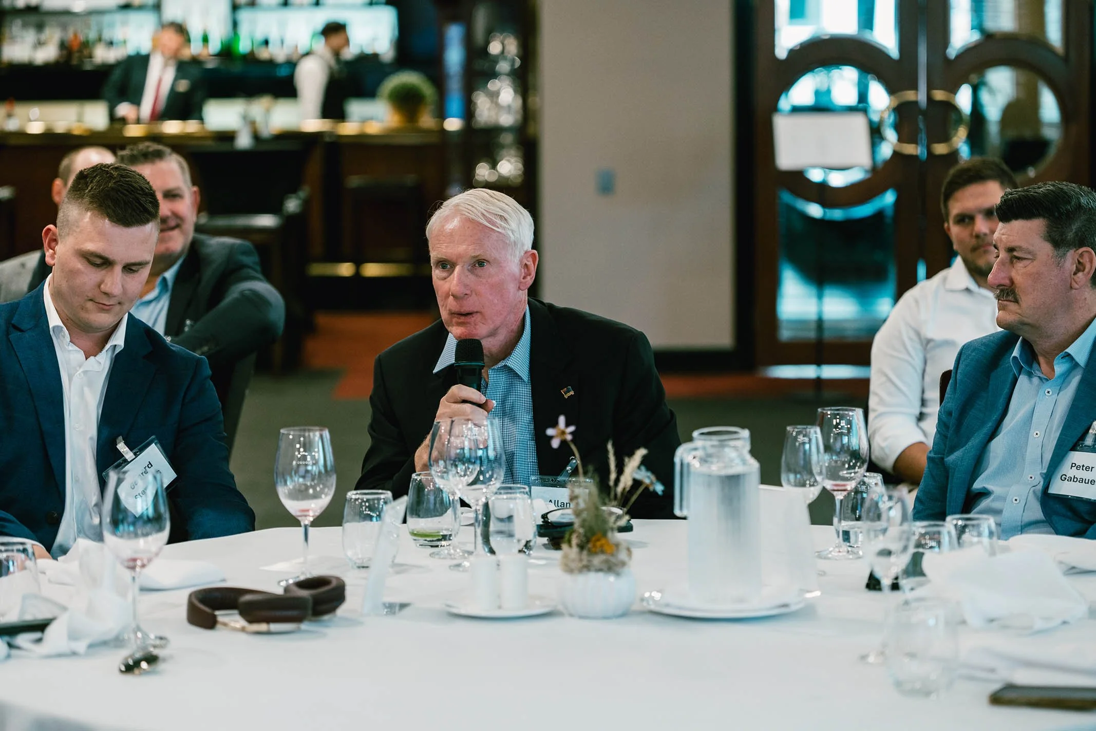 Older man holding a microphone speaking at a round table during a formal event, with several men listening attentively, in a restaurant or conference room setting.