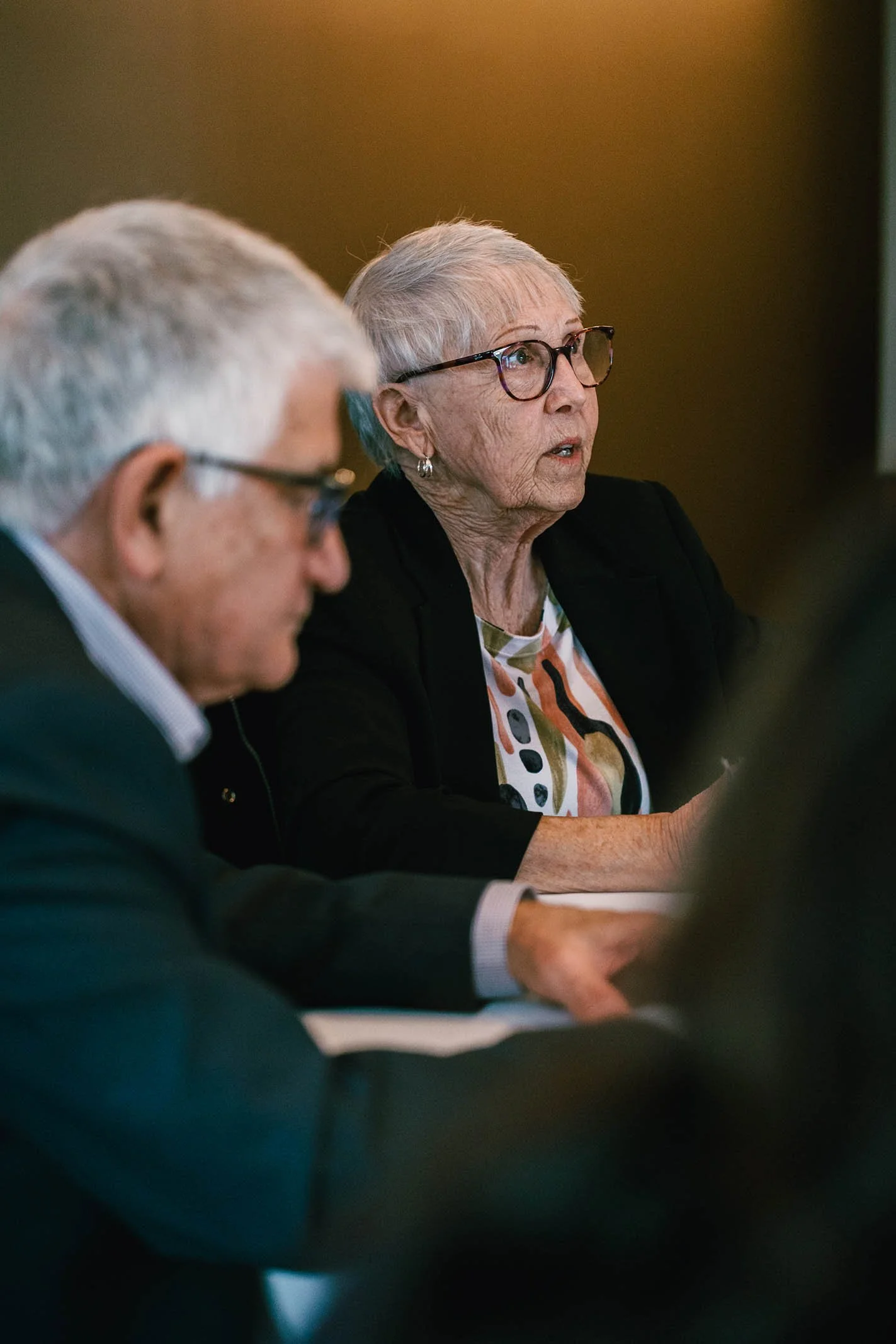 An elderly woman with short gray hair and glasses speaking or listening during a meeting, sitting at a table with a man beside her.