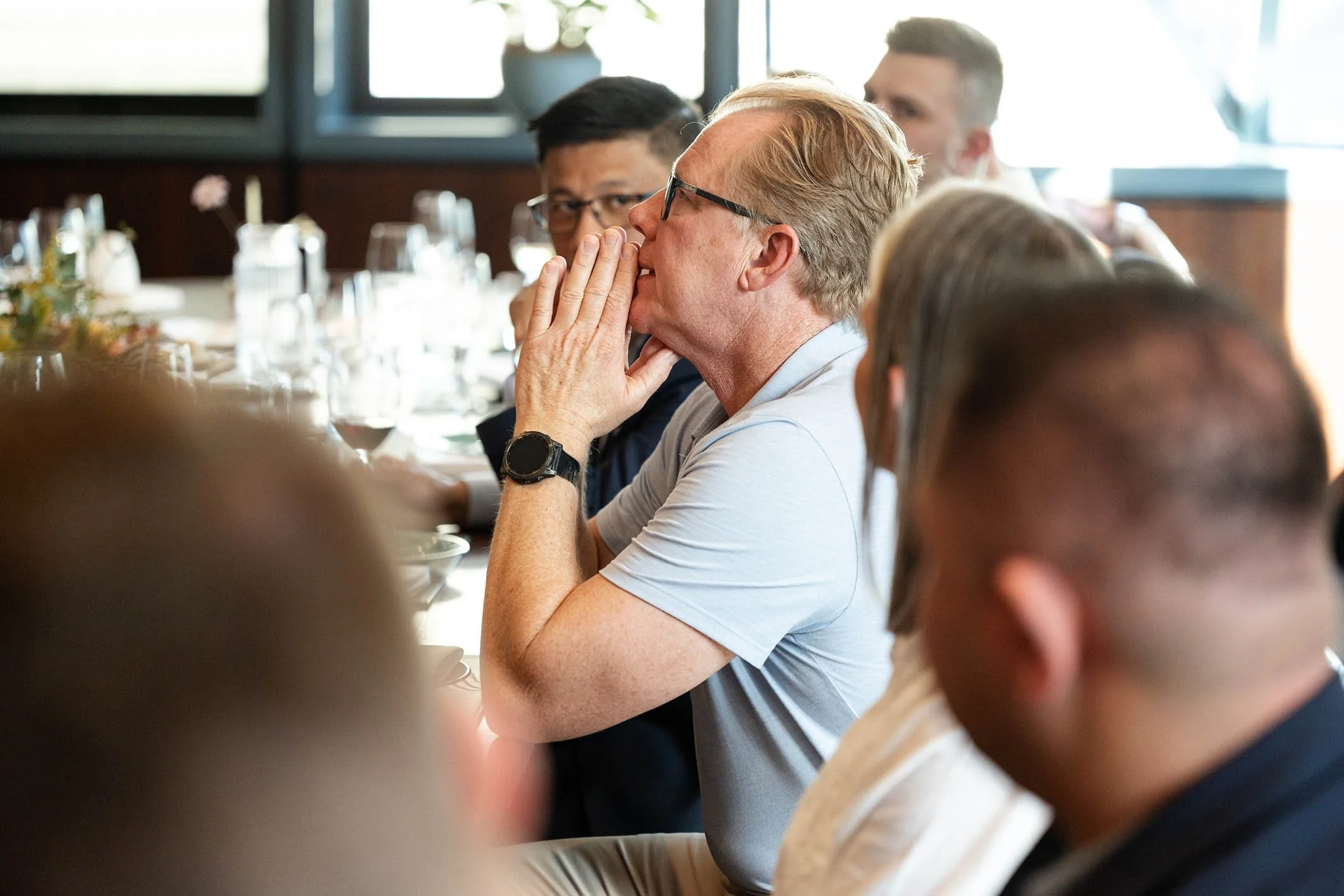 A group of people seated at a long dining table attends a formal event or meeting. The focus is on a middle-aged man with glasses, wearing a light gray shirt, with his hands folded near his face, appearing to listen attentively.