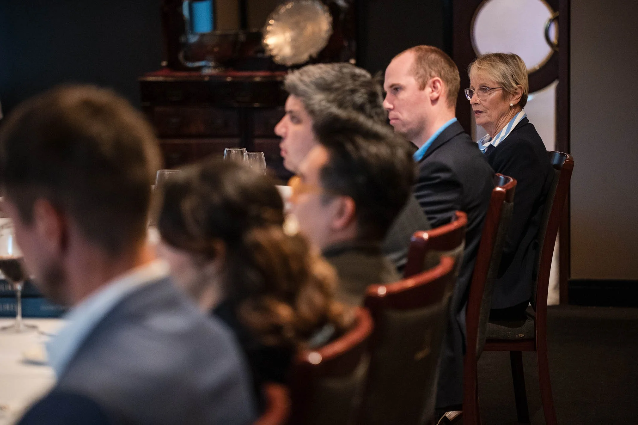 People sitting at a formal dinner event, focused on a presentation or speaker, with wine glasses on the table.