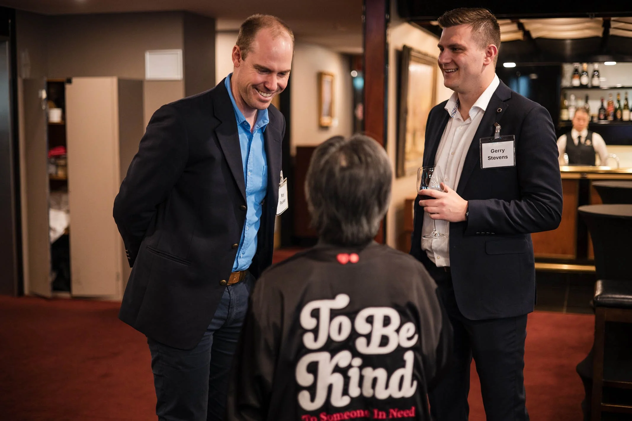 Two men in suits with name tags smiling and talking to a person in a black jacket with 'To Be Kind' written on the back at a social event in a restaurant or bar.