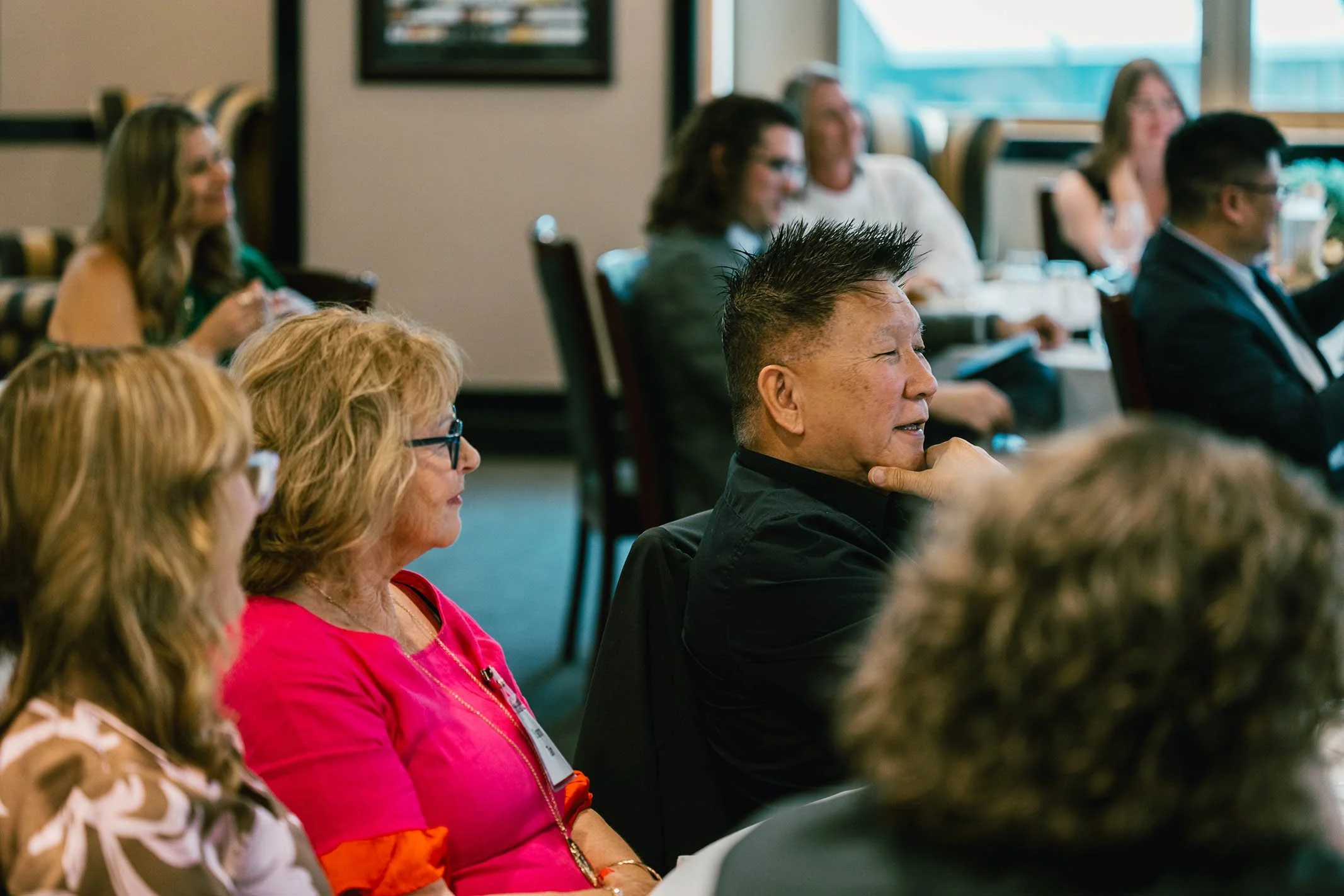 People sitting at a conference or seminar, attentively listening, in a well-lit room with large windows. In focus is a man with spiked hair, wearing a black shirt, smiling and resting his chin on his hand.