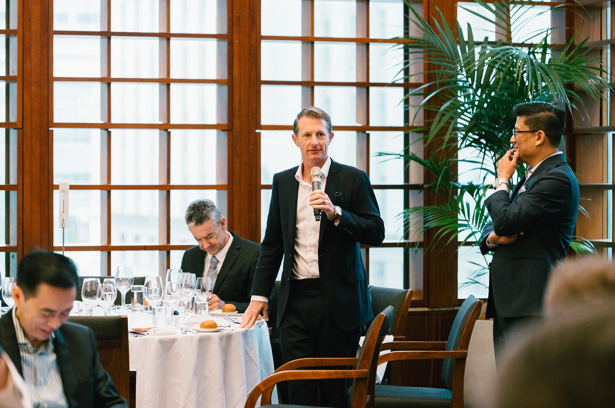 A man in a black suit holding a microphone speaking at a formal event while standing next to another man in a black suit listening. Several people are seated at tables with white tablecloths, glasses, and bread rolls inside a room with wooden window 