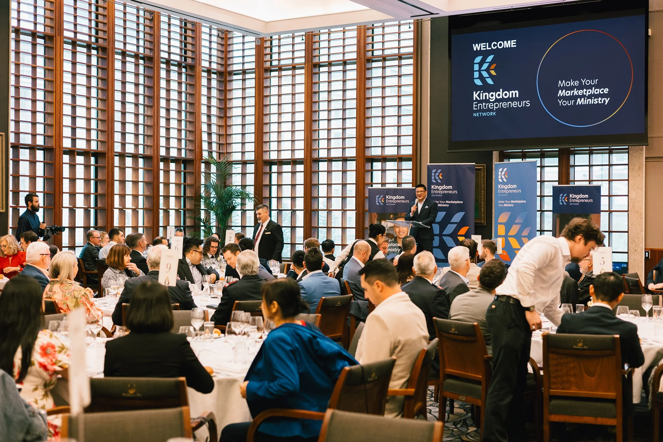 A conference room filled with attendees sitting at round tables, listening to a speaker at a podium. Large digital screen displays the Kingdom Entrepreneurs Network logo and slogan.
