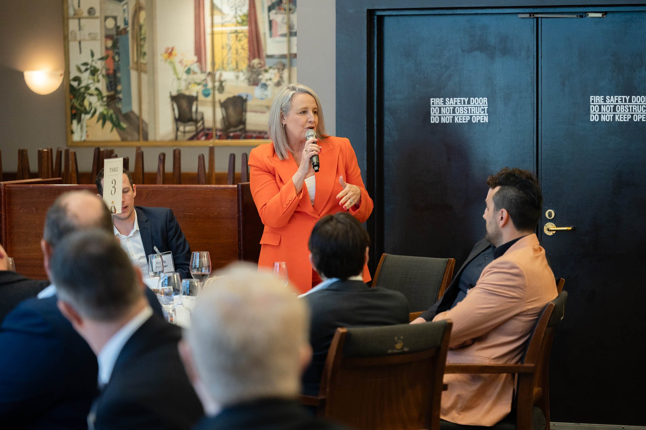 A woman in an orange blazer speaking into a microphone during a business meeting or conference, with several seated attendees listening, in a room with a large black door and a framed painting on the wall.