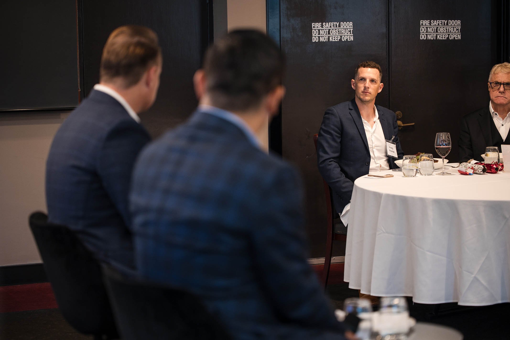 Three men in business suits sitting at a table during a formal meeting or conference, with two other men visible in the background.