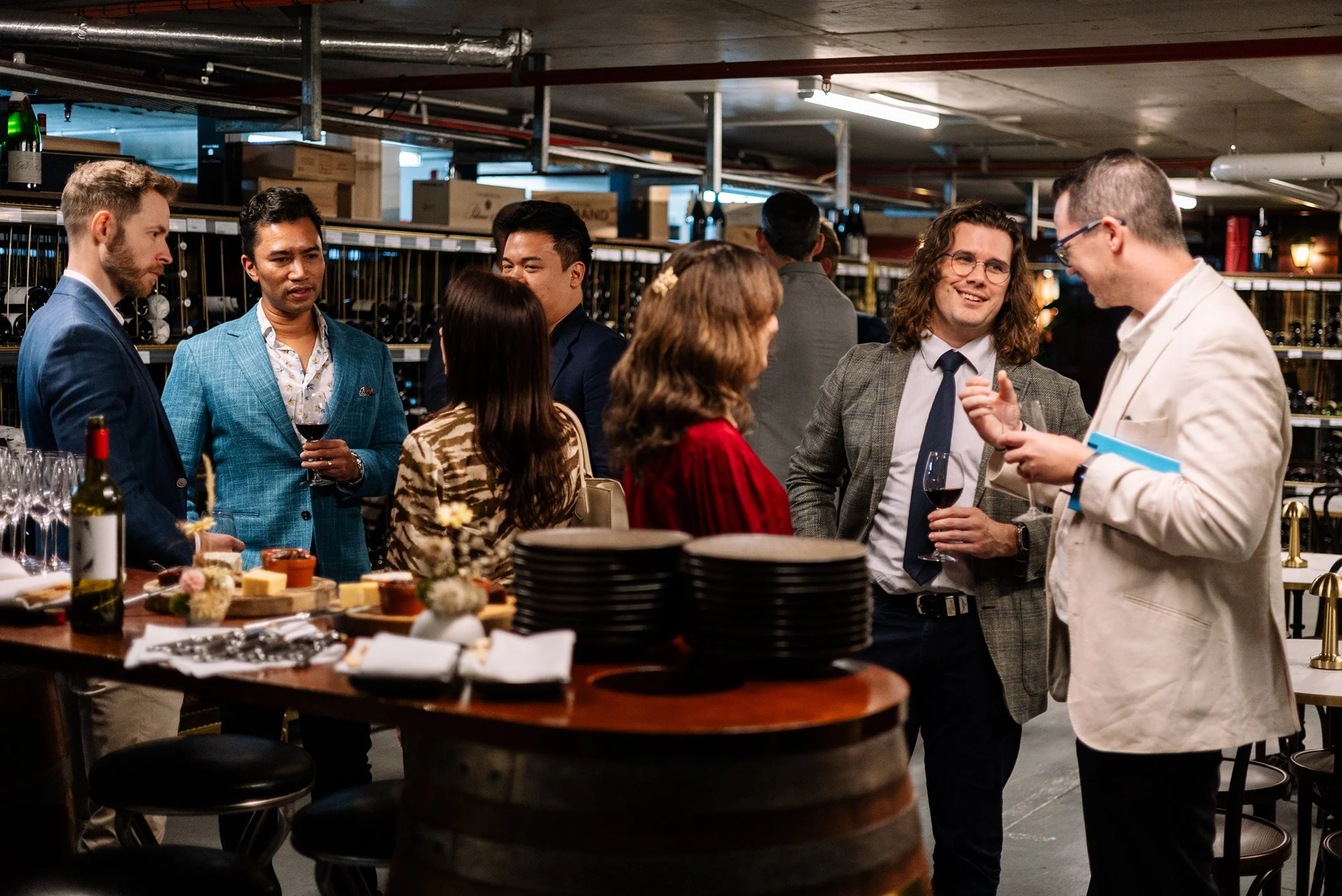 Group of people socializing at a wine tasting event, holding glasses of red wine, in a wine cellar or storage room.