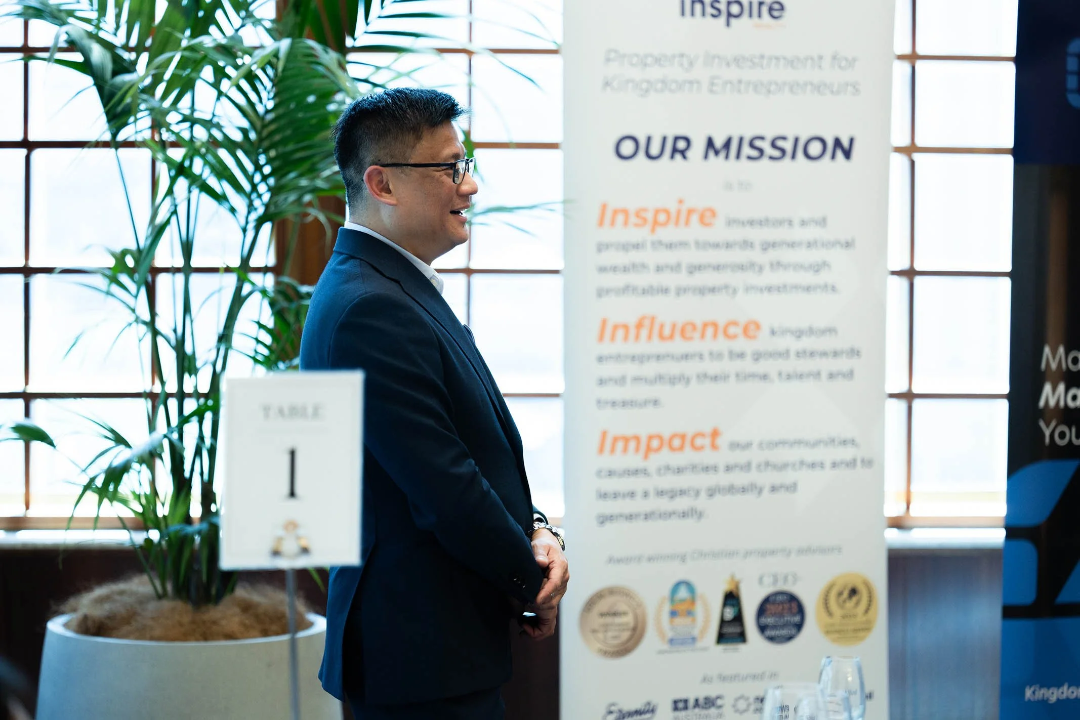 A man in a dark suit and glasses standing near a white banner with text, in a room with large windows and potted plants.