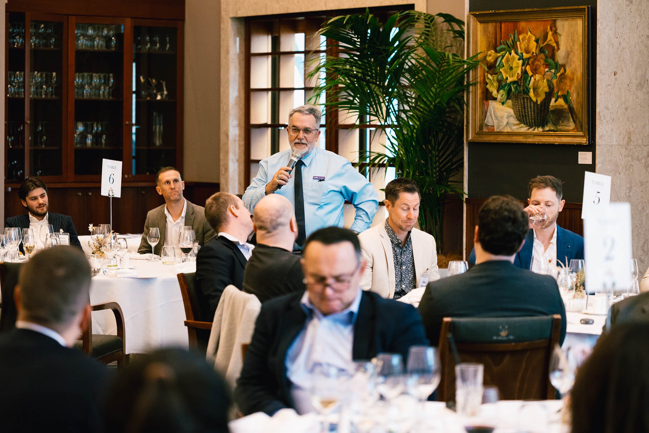 A man in a light blue shirt and dark tie speaking into a microphone at a formal dinner event, with audience members seated at round tables, some with glasses of wine, in a decorated room with artwork and large plant.