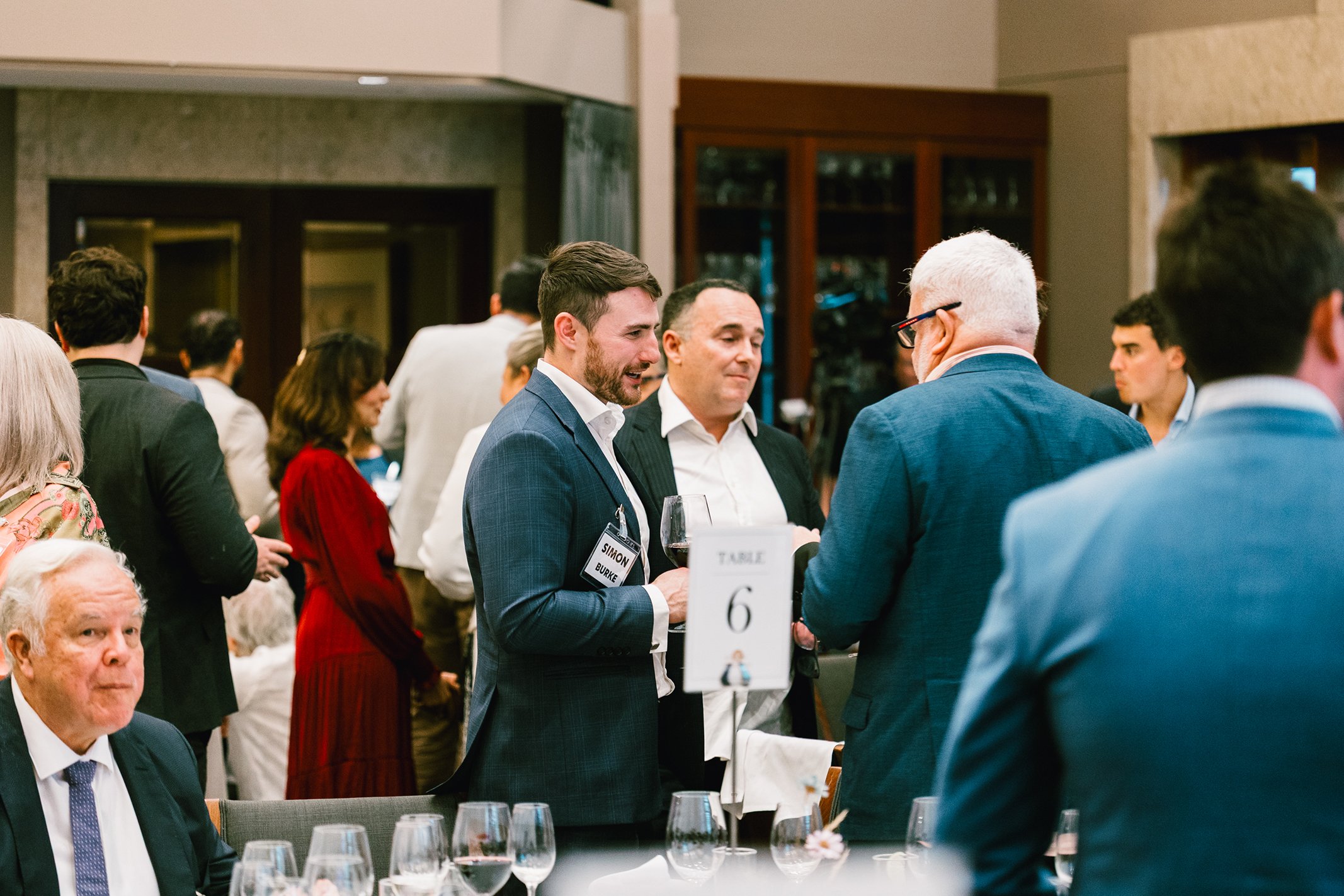 People attending a formal networking event or conference, conversing and holding drinks around a table.