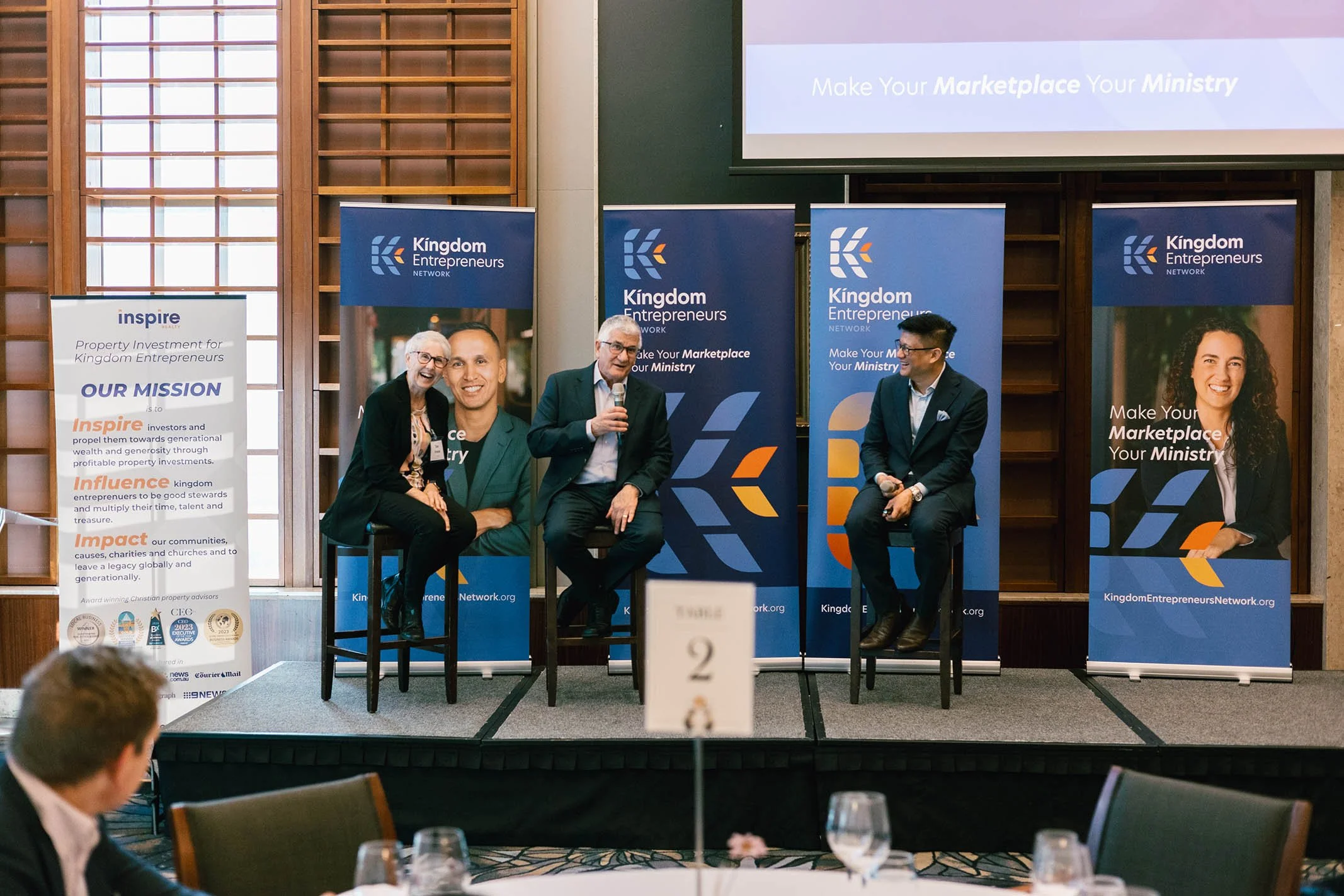 A panel discussion at a Kingdom Entrepreneurs event with four individuals on stage, sitting on stools with banners behind them, including a woman on the left, a man with glasses holding a microphone, another man with glasses, and a woman on the right