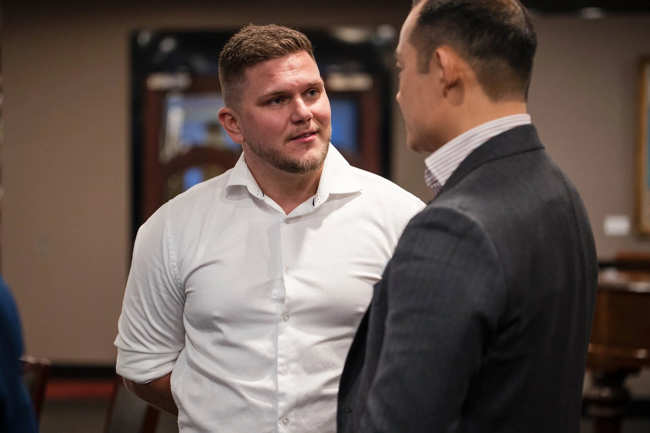 Two men are engaged in a conversation indoors, one with short light brown hair and a beard wearing a white shirt, and the other with dark hair wearing a dark blazer and a striped shirt.
