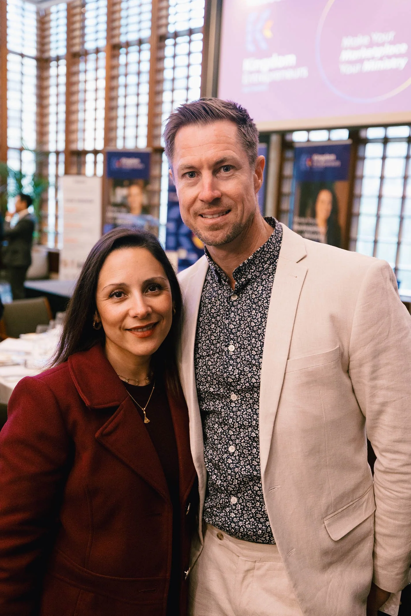 A woman and a man standing close together at an indoor event, smiling for the camera, with a blurred background featuring banners and people.