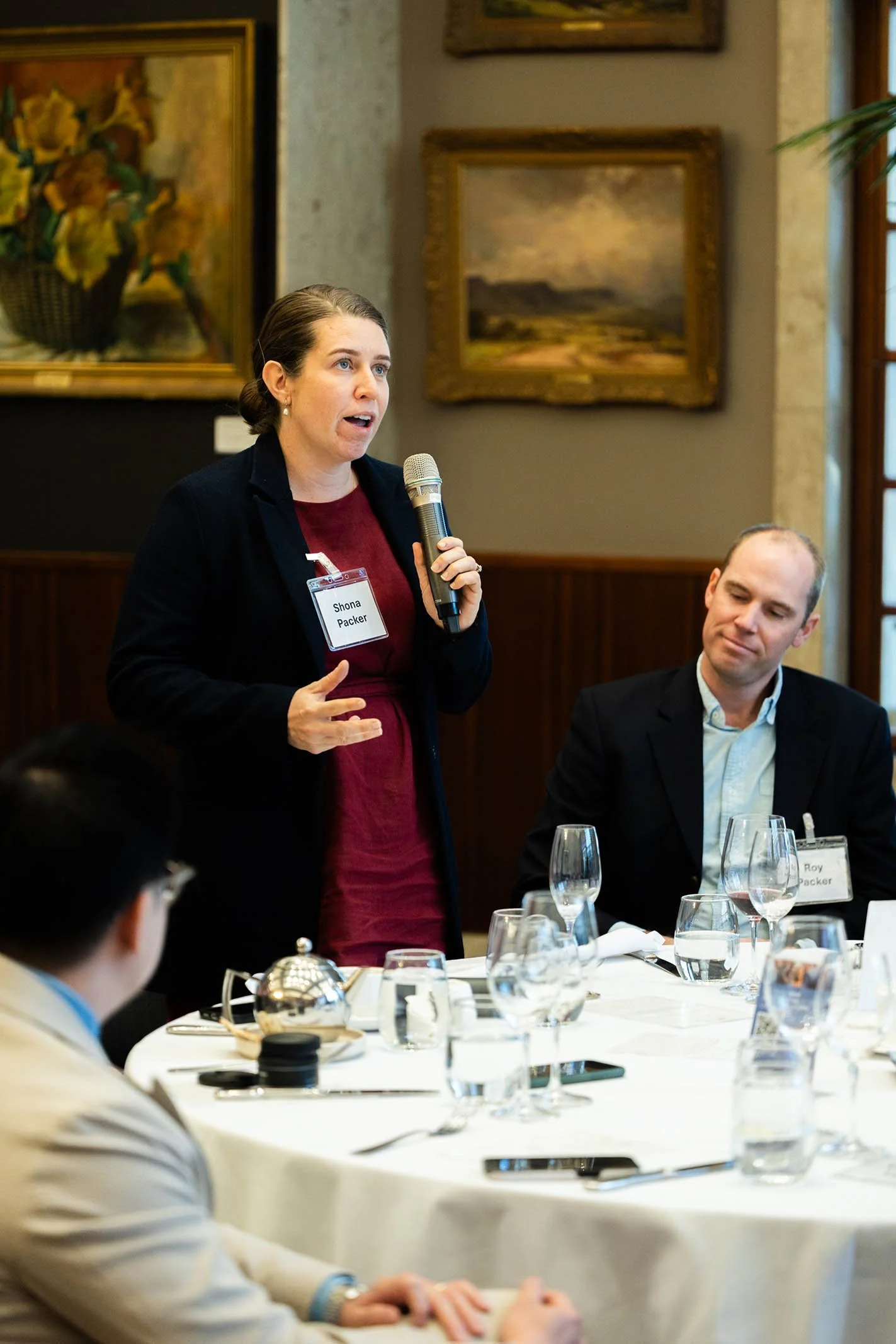 A woman standing and speaking into a microphone during a formal meeting or conference, with name tag 'Shona Packer'. Seated man looks at her, surrounded by a round table with glasses, plates, and a teapot. Artwork decorates the wall behind them.