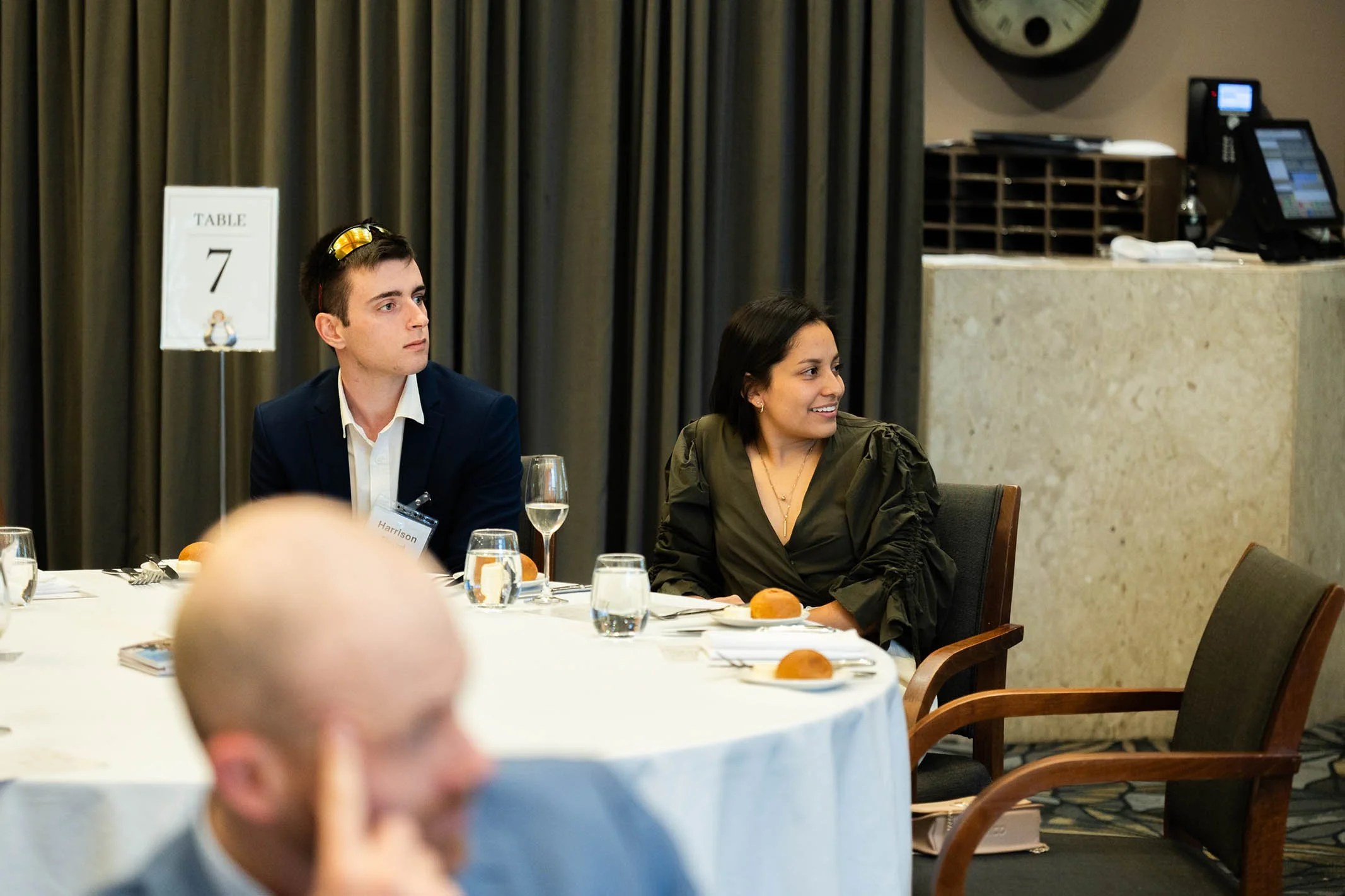 Two people sitting at a dinner table with wine glasses and bread rolls, attending a formal event in a hotel conference room identified by a table sign labeled '7'.