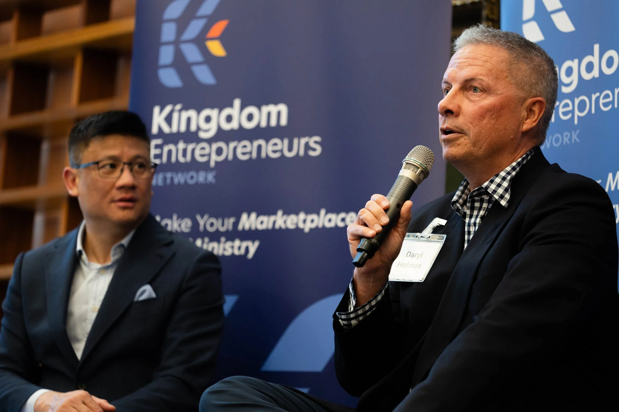 Two men sitting on a stage at a Kingdom Entrepreneurs event, with one speaking into a microphone. A blue banner with the event logo and text is visible in the background.