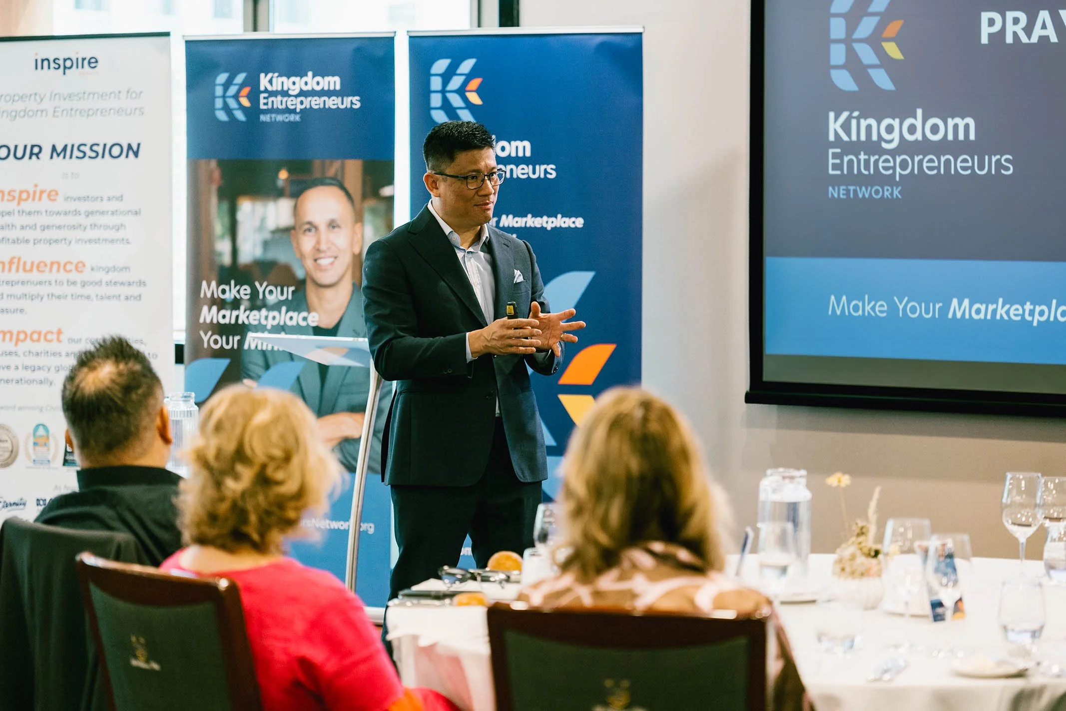 Man in suit giving presentation at Kingdom Entrepreneurs Network event, with a screen and banners in the background, and audience seated at table.