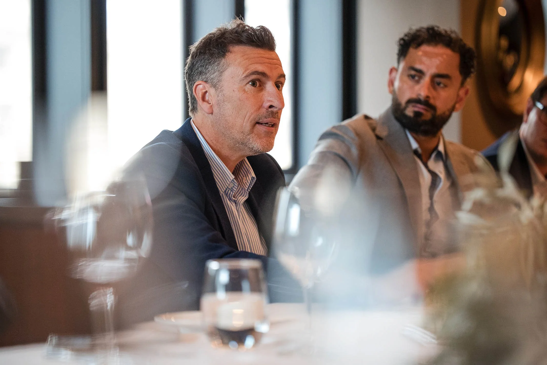 Two men are sitting at a table during a meeting or conference. One man has gray hair, a beard, and is wearing a dark blazer and a striped shirt. The other man has dark curly hair, a beard, and is wearing a light gray blazer and a shirt. There are gla