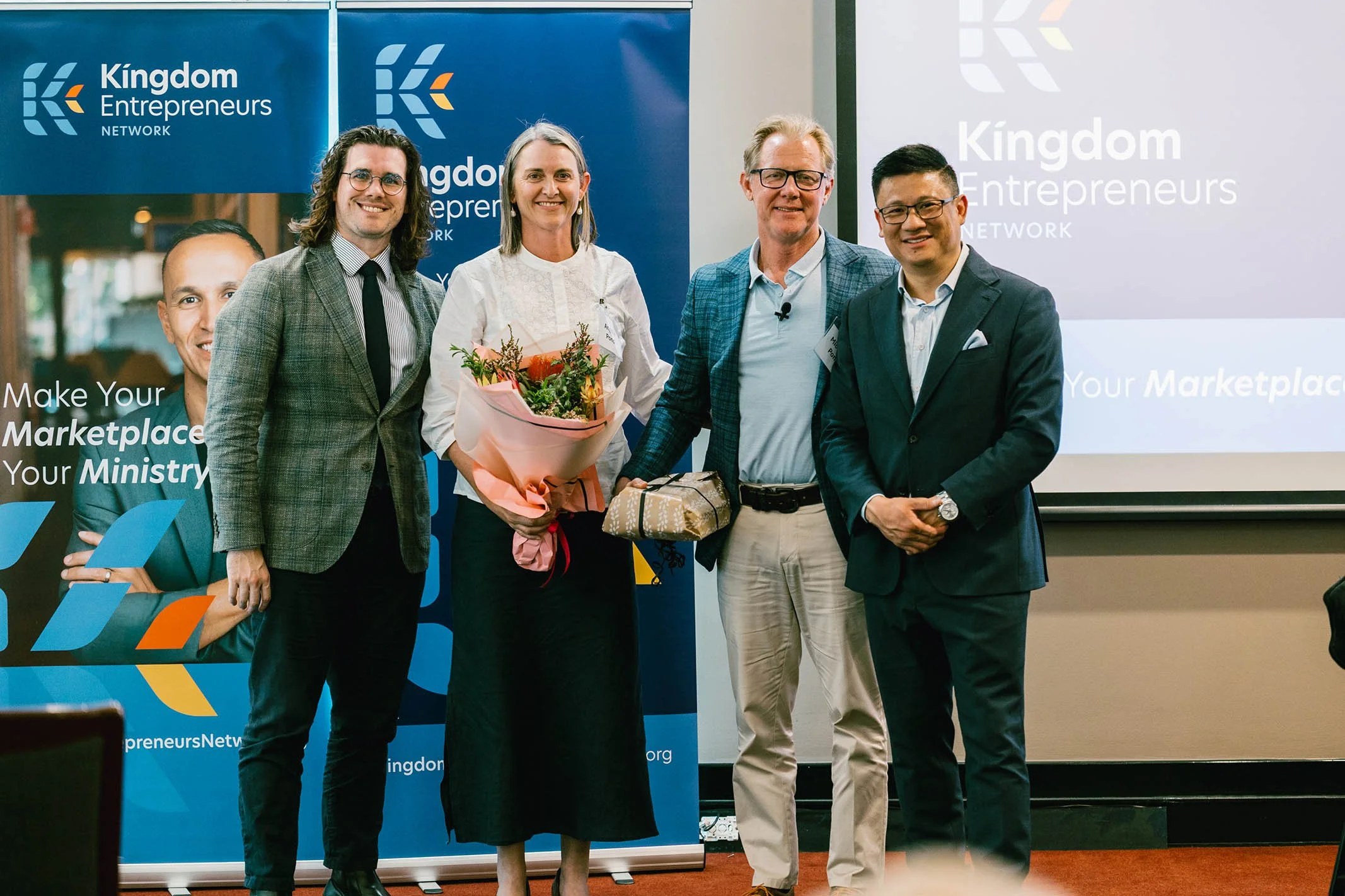 Five people standing in front of a blue banner and a projection screen, with one woman holding a bouquet of flowers and receiving a gift. They are dressed in business attire and smiling at the camera.