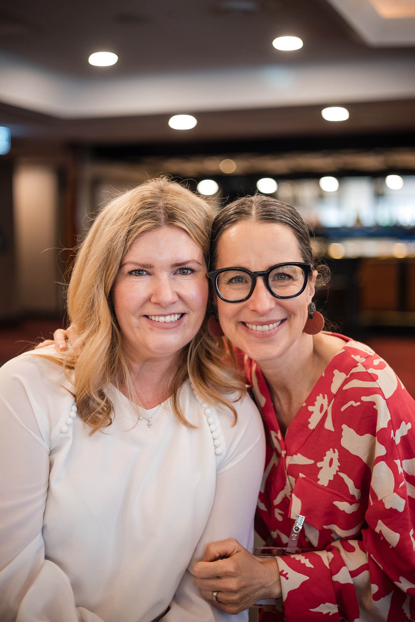 Two women smiling and posing together indoors, one with blonde hair wearing a white top and the other with dark hair, glasses, and a red and white patterned top.
