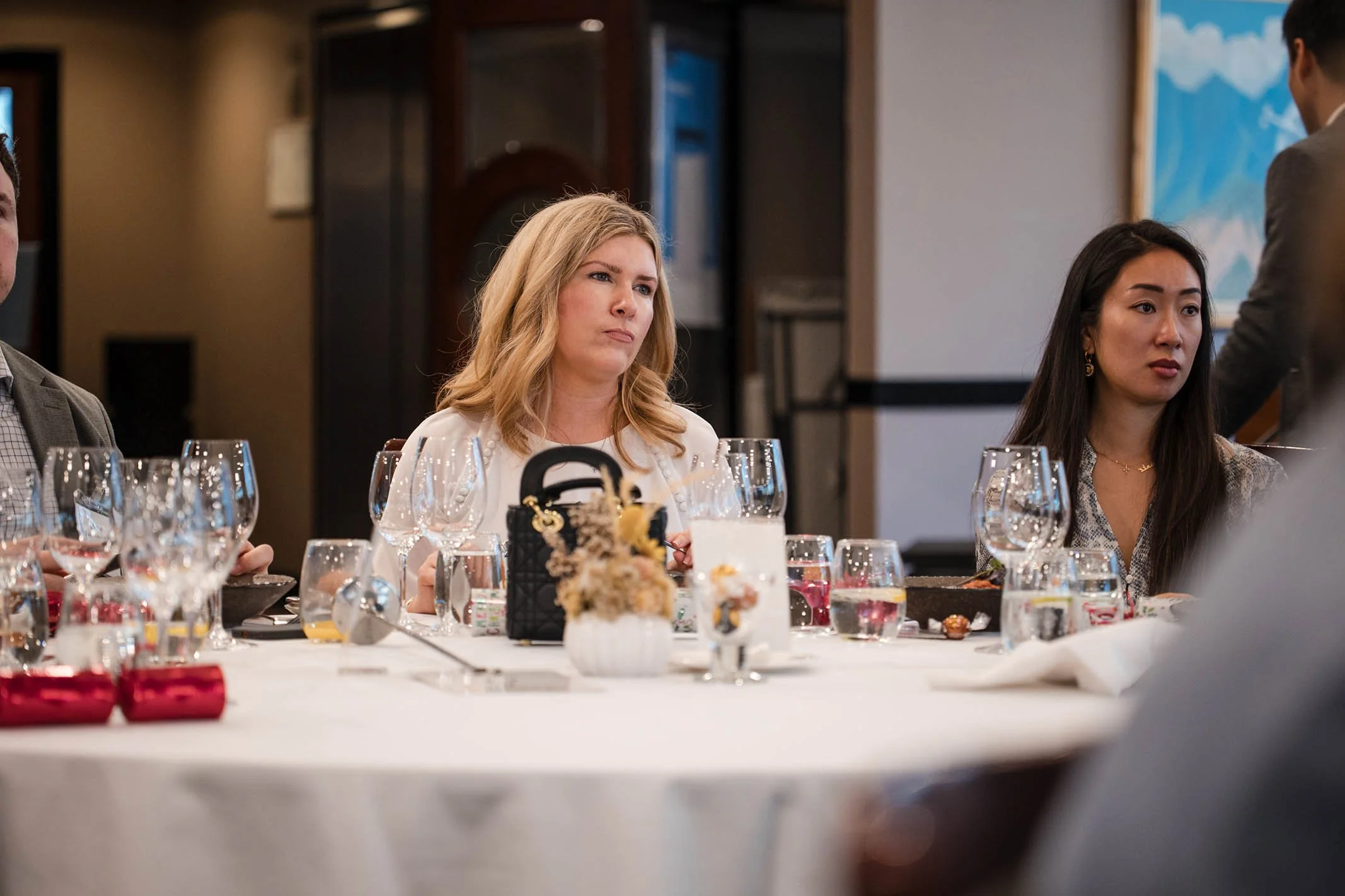 People sitting at a dinner table with wine glasses and floral centerpieces, showing a woman with blonde hair and serious expression in the center and a woman with dark hair on the right.