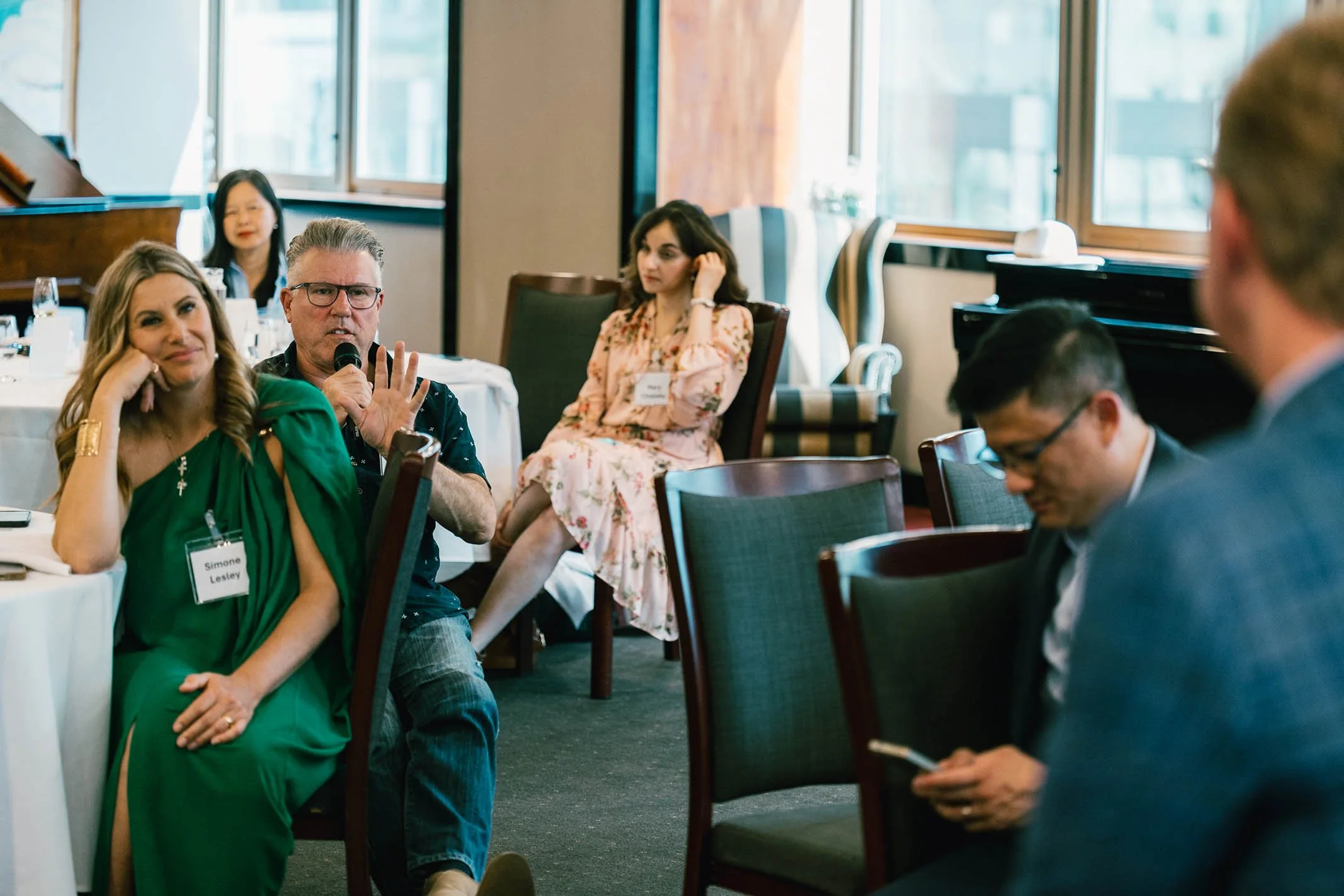 A woman in a green dress with a name tag sitting at a table, a man next to her holding a microphone and speaking, and other people seated around in a conference room with large windows.