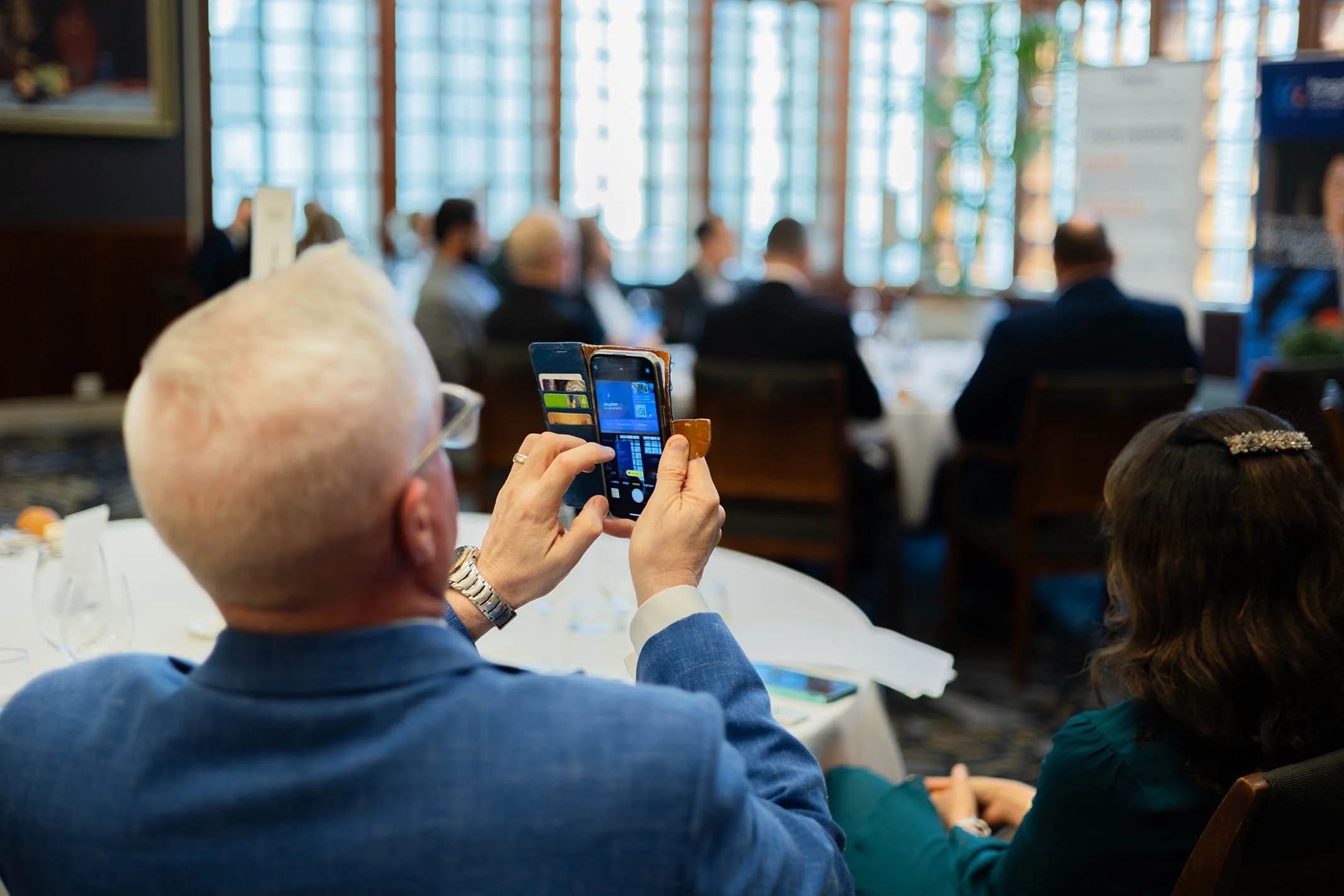 An elderly man with white hair, wearing glasses and a blue suit, is seated at a dining table, taking a photo with his smartphone during a formal event in a well-lit room with large windows and other people seated at tables in the background.
