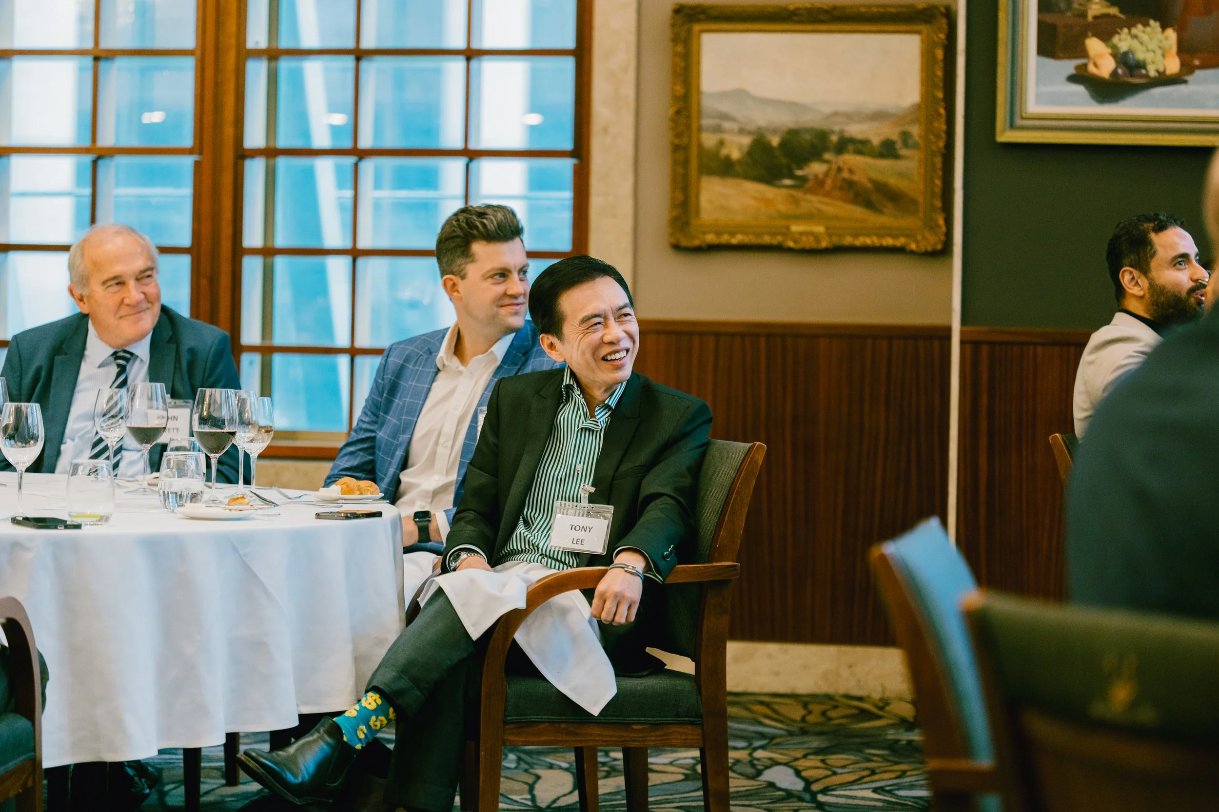 A group of professionally dressed men seated at a round table in a conference or banquet room, with wine glasses and plates on the table, laughing and smiling during a formal event.