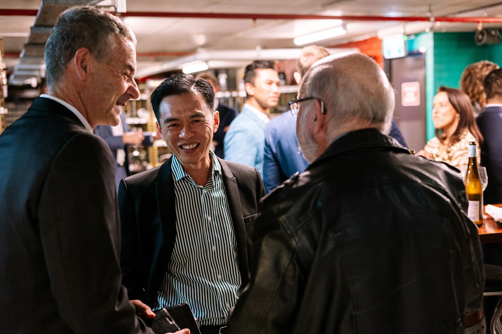 Three men in conversation at a lively social gathering, smiling and dressed in formal and casual attire, with other guests in the background.