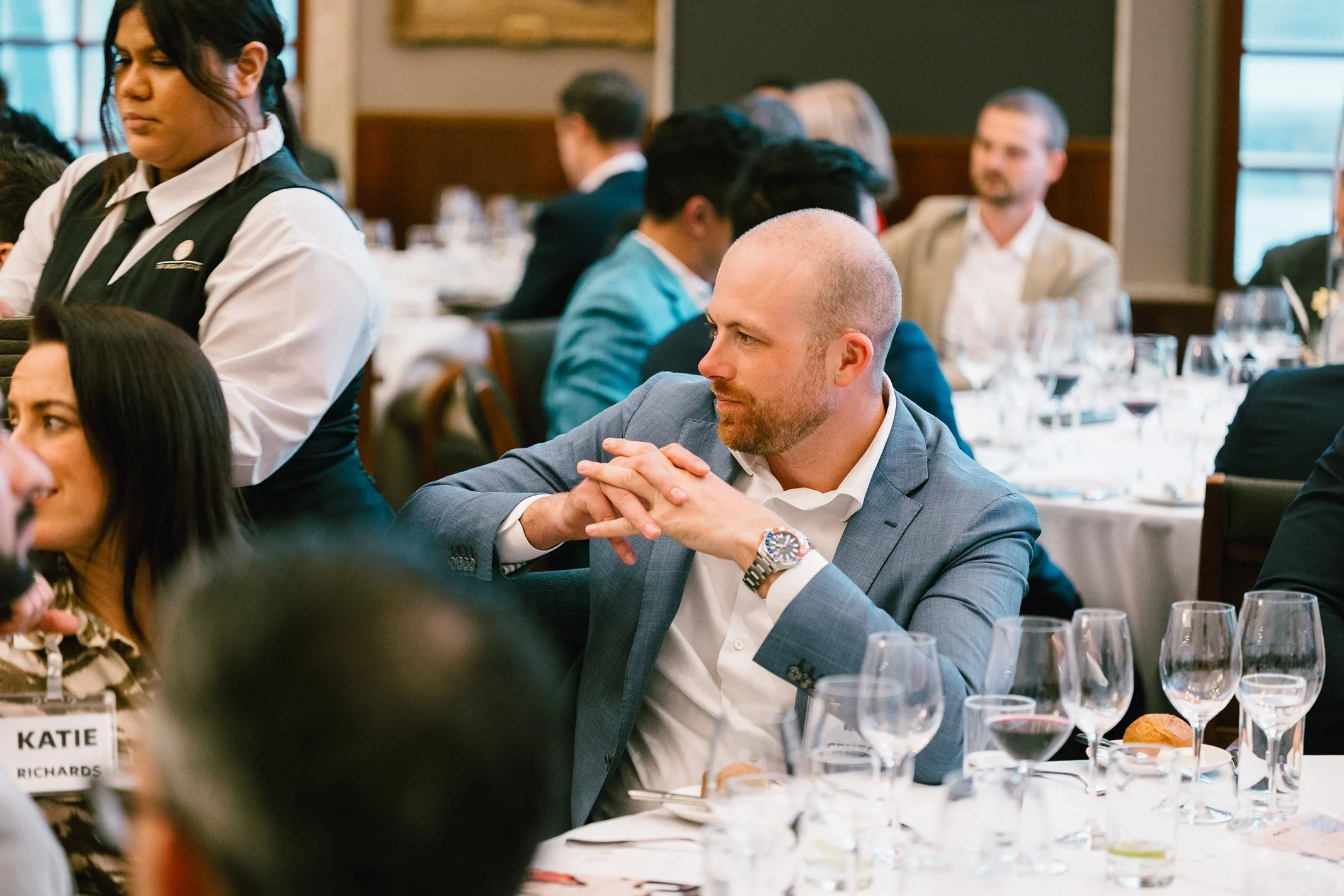 A man in a gray suit and white shirt sitting at a formal dining table with his hands clasped, //enjoying a meal at a formal event, surrounded by other guests and wine glasses.