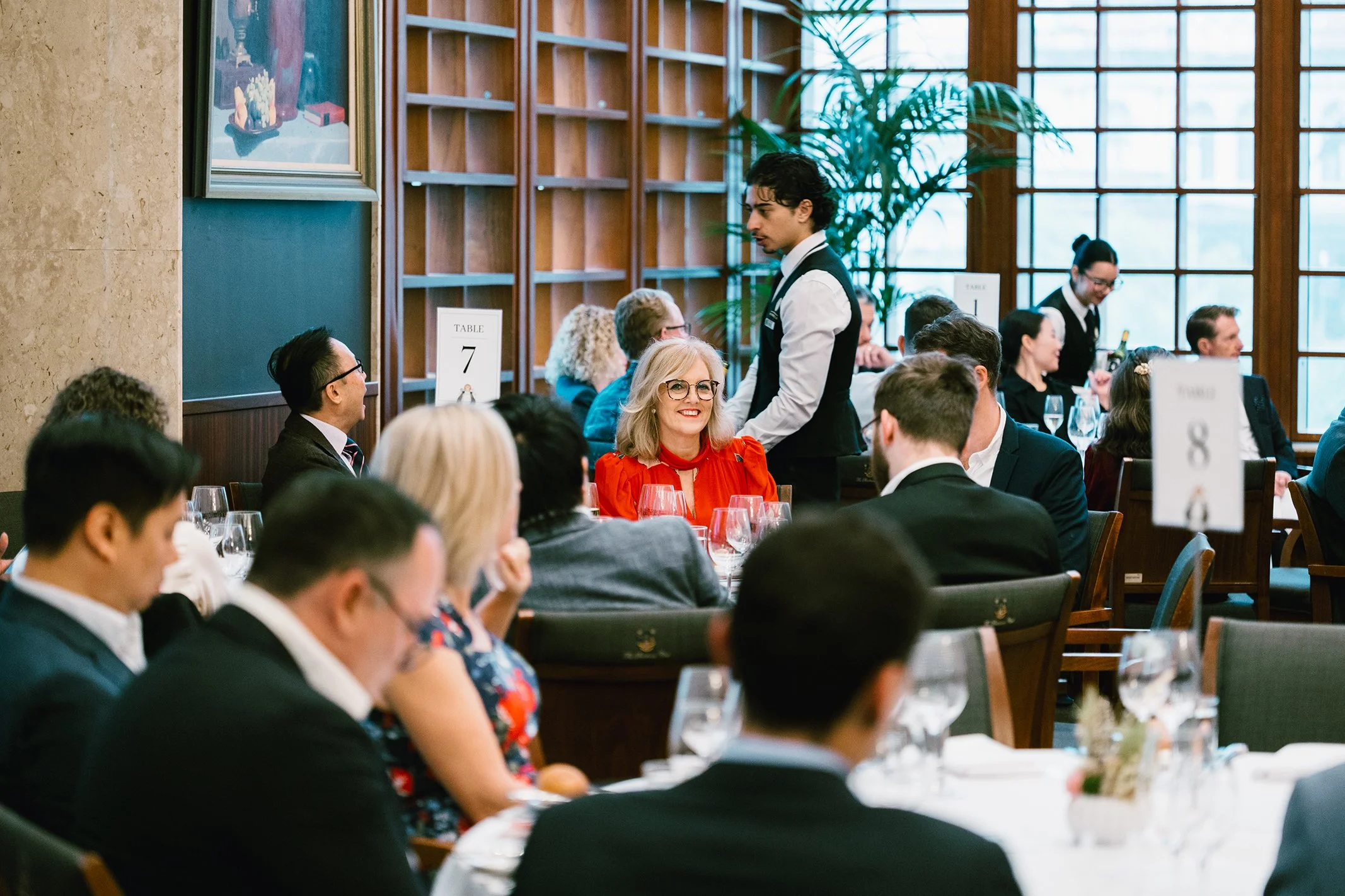 A group of people seated at a formal dining event in a well-lit room with large windows, a woman in a red dress smiling at a waiter, other guests engaged in conversations, and dining tables set with wine glasses and silverware.