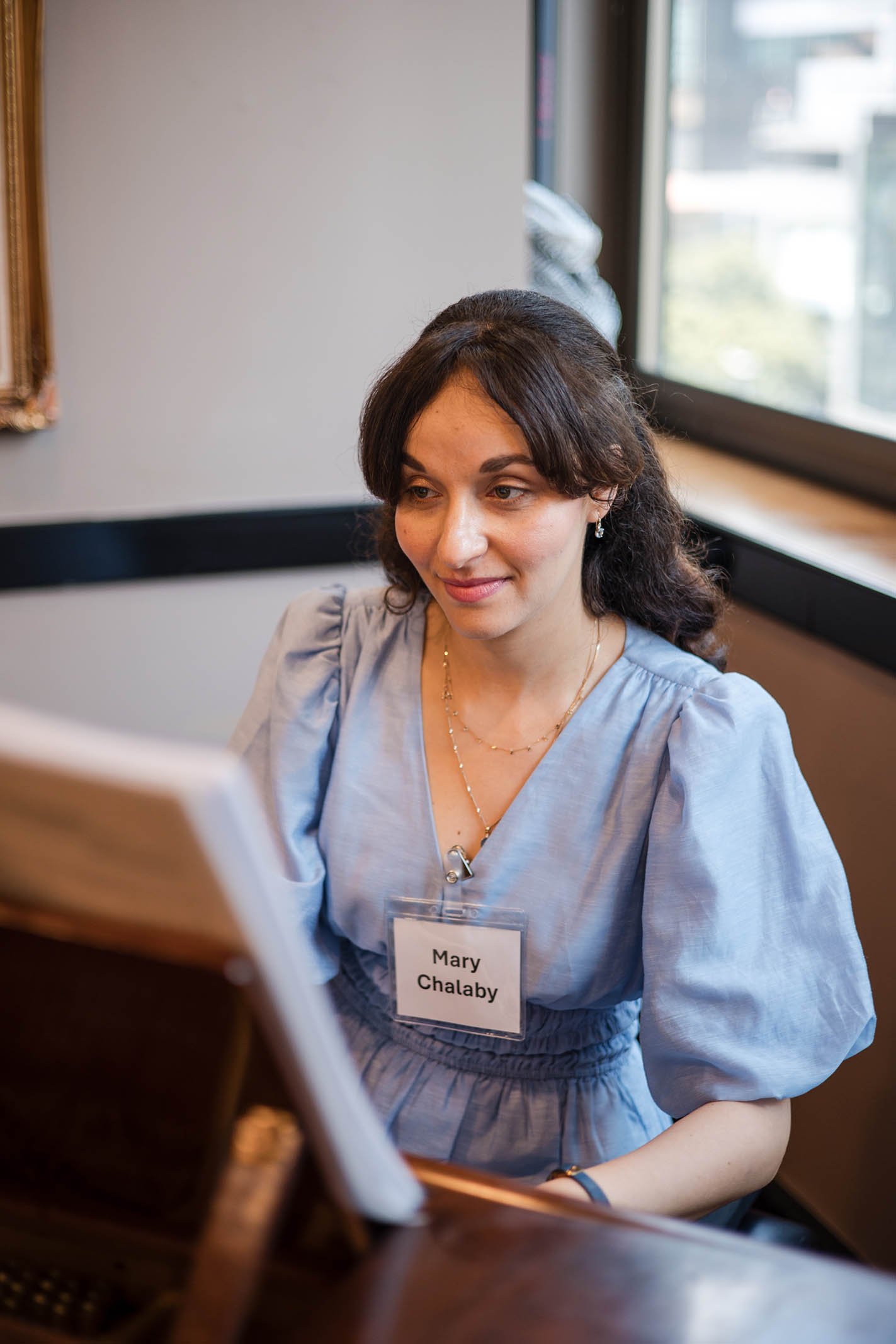 A woman with dark brown hair sits at a table, looking at a screen, wearing a light blue dress with puffy sleeves and a name tag that reads 'Mary Chalaby'. She is in a room with a window showing a city view outside.