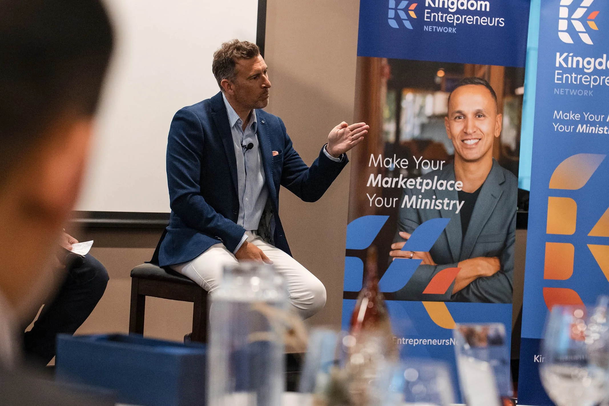 A man in a blue blazer and white pants sitting on a stool, speaking with hand gestures at a professional event. Behind him, a banner displays the logo and branding for Kingdom Entrepreneurs Network, featuring a smiling man in a grey blazer. In the fo